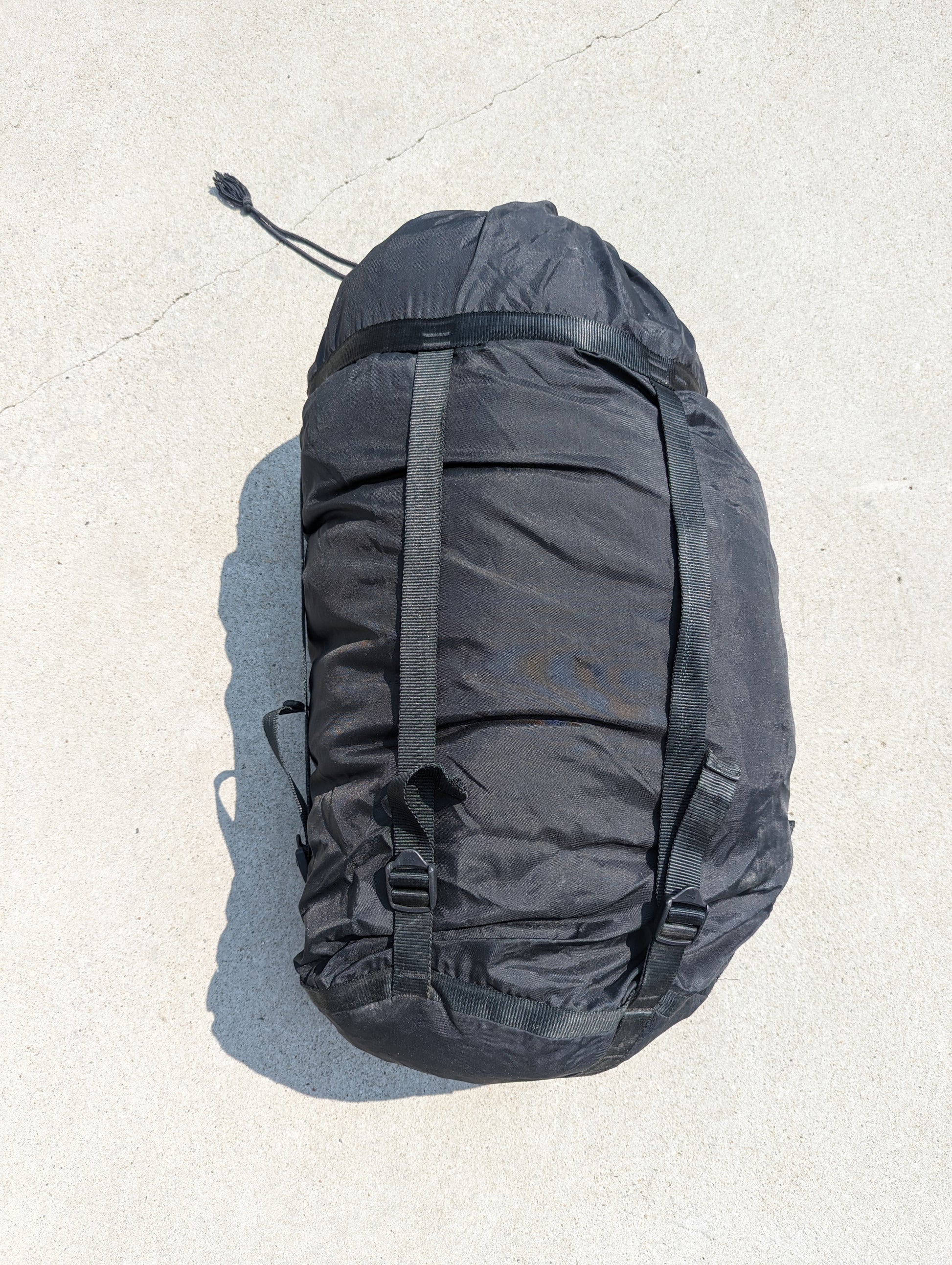 A large, black U.S. Military Surplus Compression Stuff Sack, tightly compressed into a cylinder by black webbing straps, highlighting the wrinkled nylon texture on a concrete background.