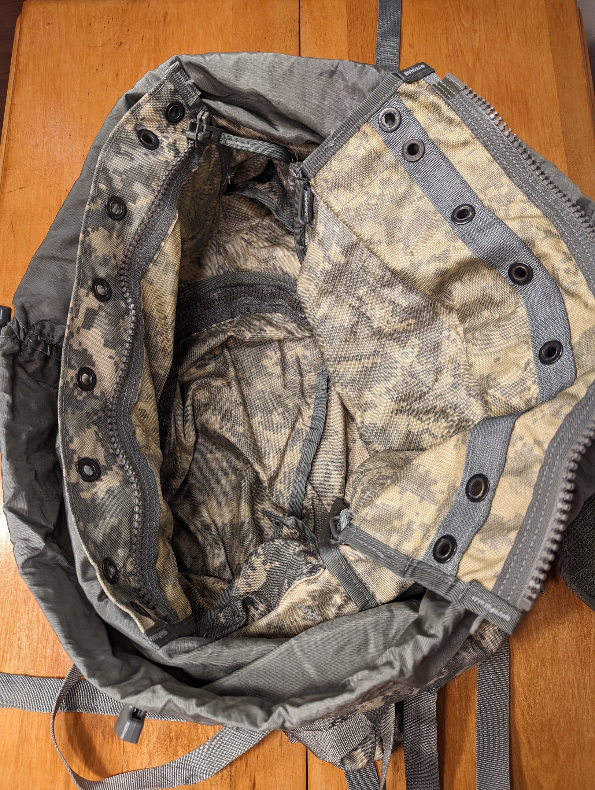 A close-up, top-down view looking into the open main compartment of a U.S. Military Large Field Rucksack in ACU camouflage, highlighting the drawstring closure, black metal grommets around the rim, and the internal zippered pocket.