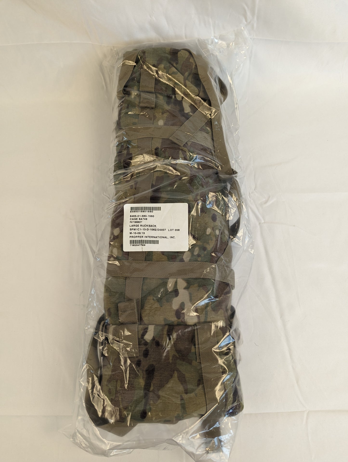 A large U.S. Military Surplus OCP/Multicam Rucksack main bag, tightly rolled and sealed inside a clear plastic bag, with compression straps and an identification tag visible.