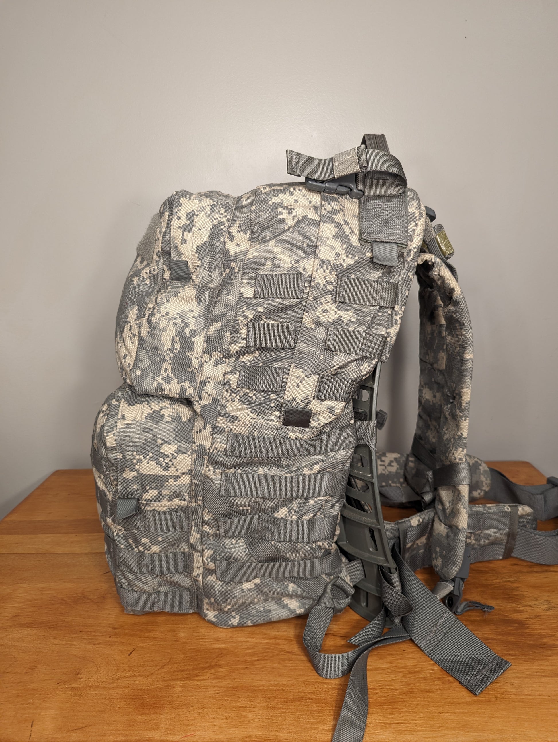 A side view of a U.S. military surplus medium field pack in ACU/UCP camouflage pattern, standing on a wooden table and featuring extensive MOLLE webbing, multiple pockets, and padded shoulder straps.