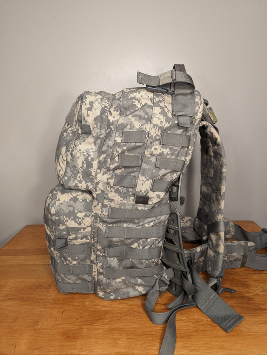 A side view of a U.S. military surplus medium field pack in ACU/UCP camouflage pattern, standing on a wooden table and featuring extensive MOLLE webbing, multiple pockets, and padded shoulder straps.