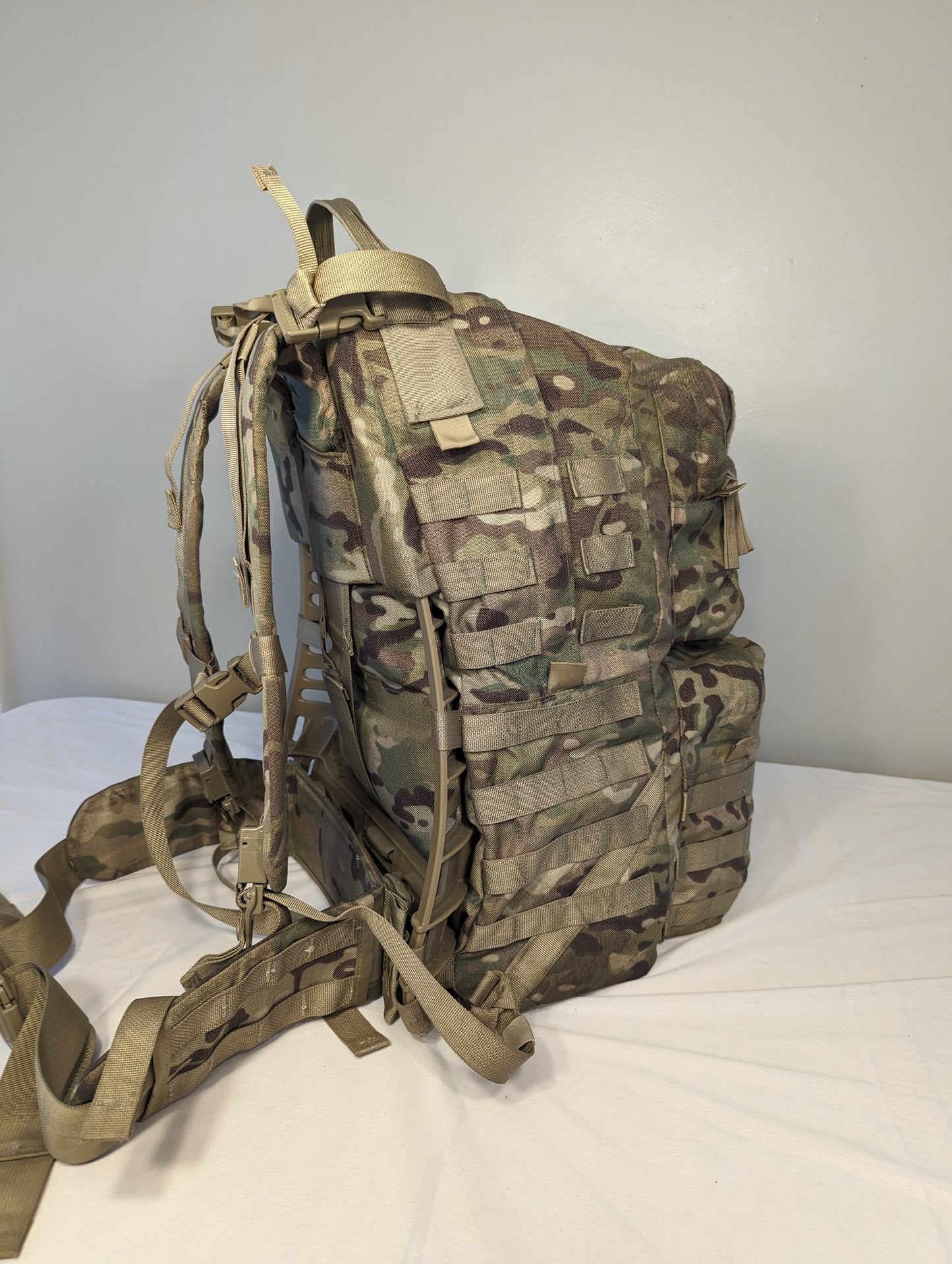 A side profile view of a U.S. Military Surplus Medium Field Pack Rucksack in OCP/Multicam camouflage, standing upright, highlighting the bulky profile, horizontal MOLLE webbing, and padded shoulder harness.