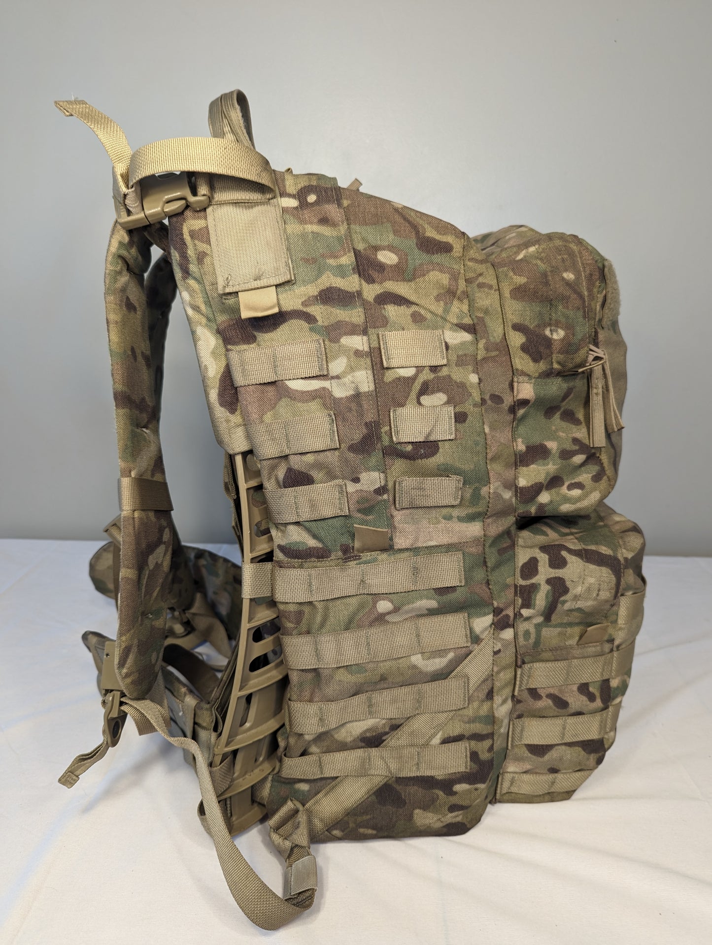 A side profile view of a U.S. Military Surplus Medium Field Pack Rucksack in OCP/Multicam camouflage, standing upright, highlighting the bulky profile, extensive horizontal MOLLE webbing, and side compression straps.