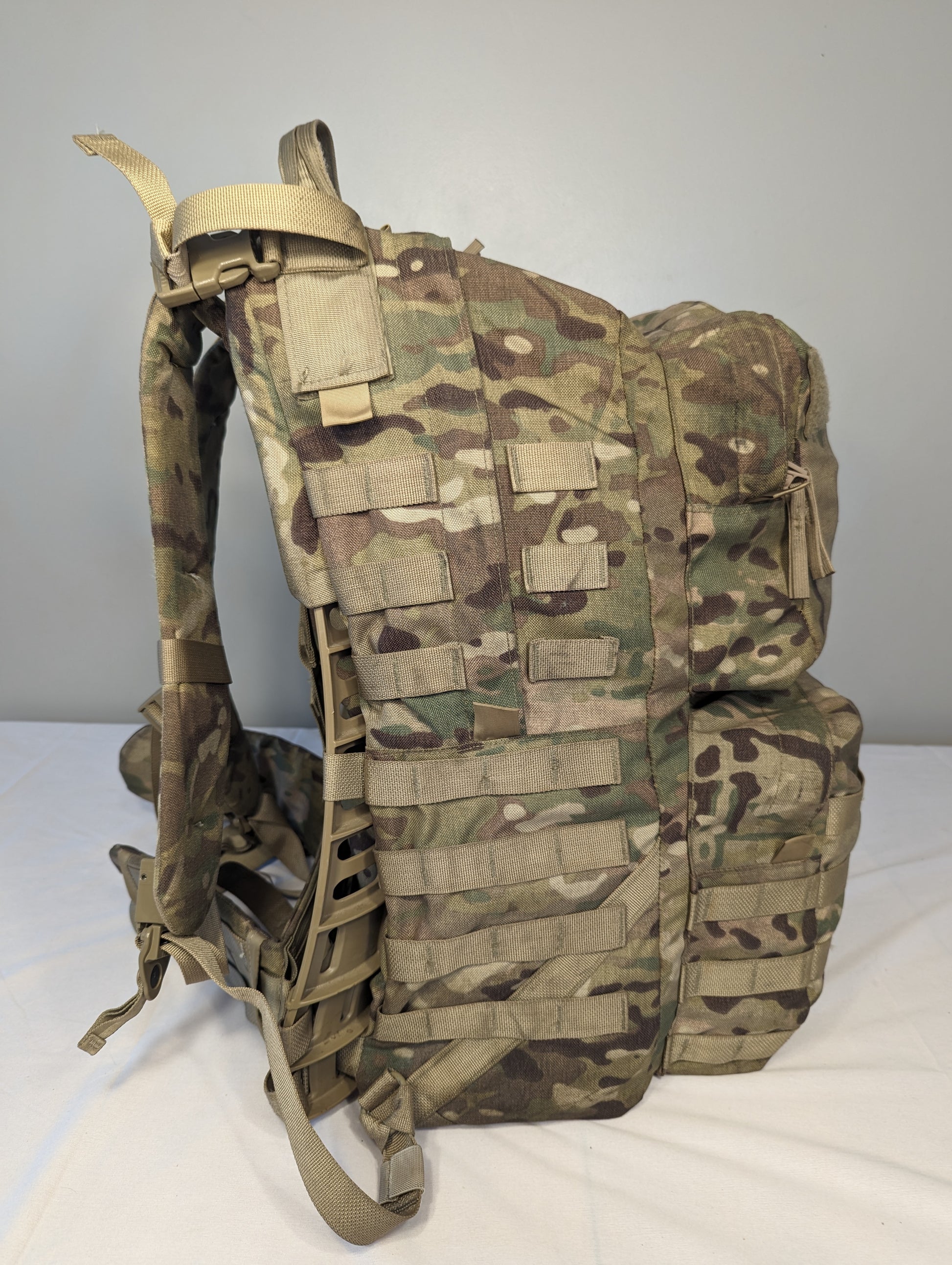 A side profile view of a U.S. Military Surplus Medium Field Pack Rucksack in OCP/Multicam camouflage, standing upright, highlighting the bulky profile, extensive horizontal MOLLE webbing, and side compression straps.