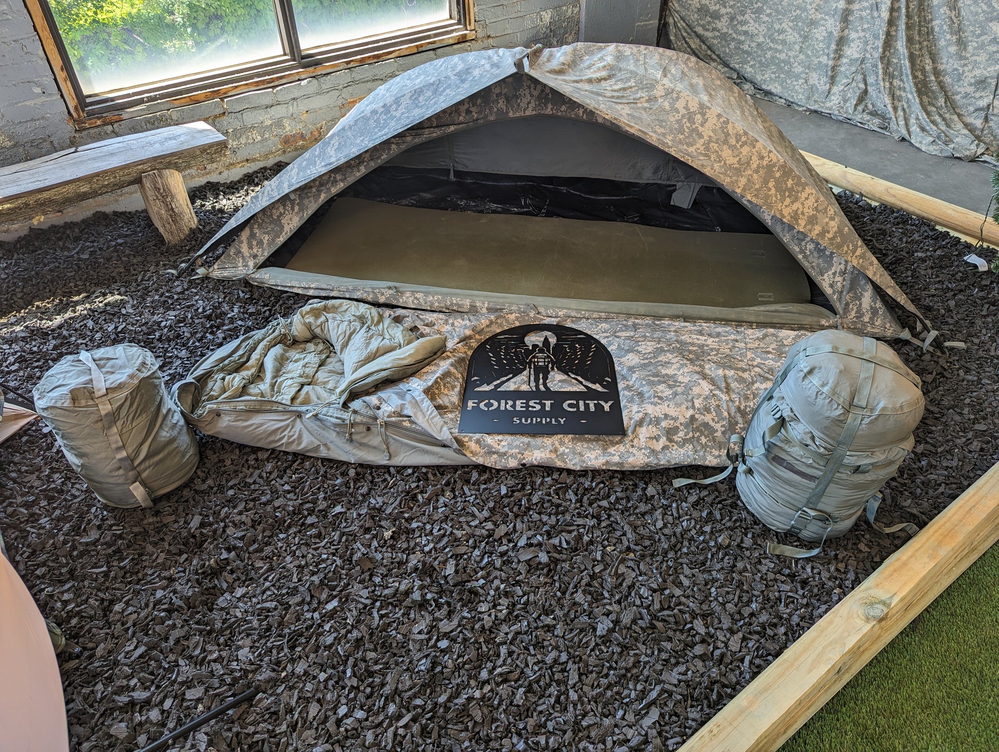 A pitched U.S. Military Modular Sleep System Bivy Cover in ACU camouflage, set up as a one-person tent on a dark mulch floor, with two gear sacks and the 'FOREST CITY SUPPLY' logo resting at the entrance.