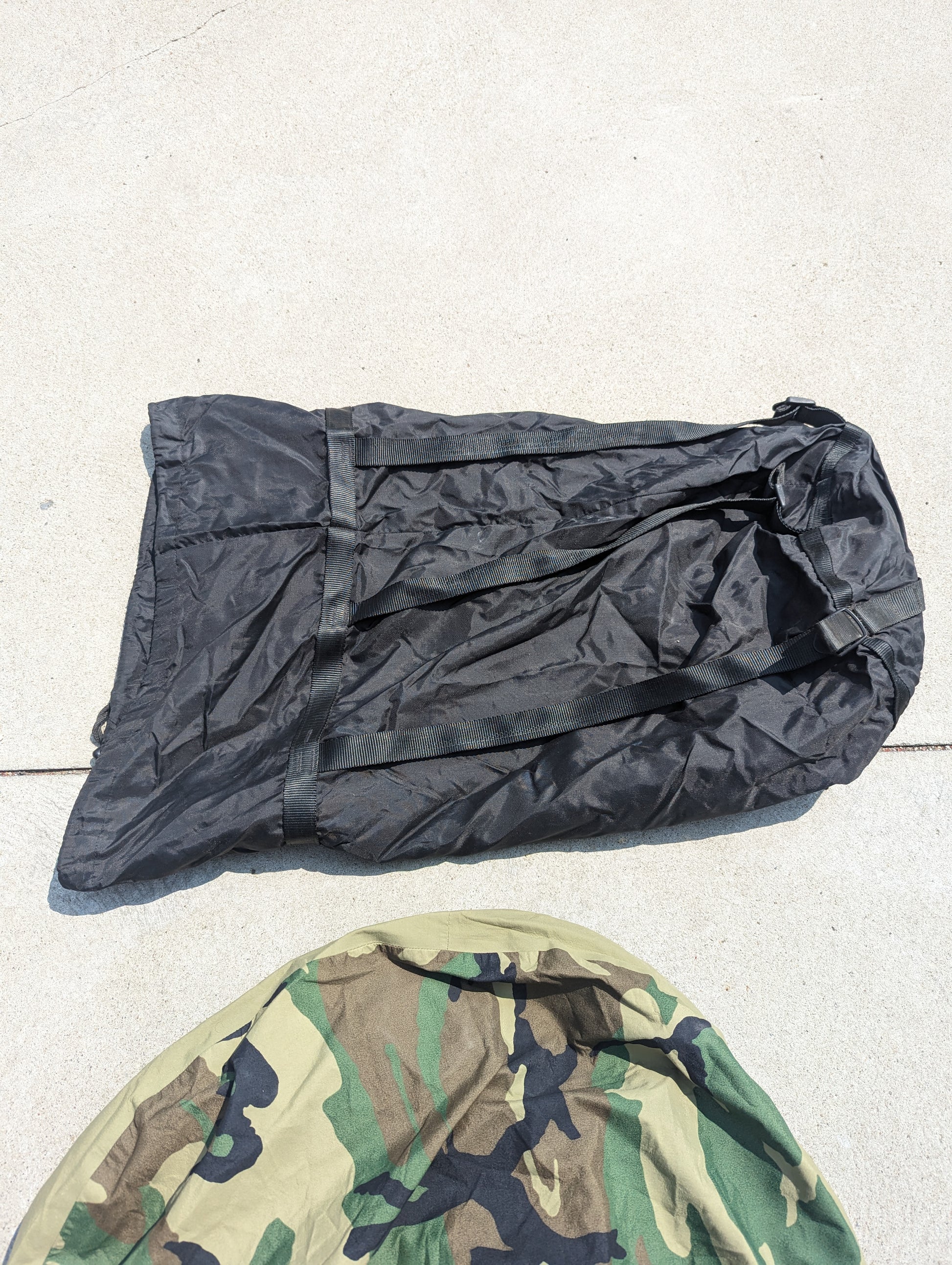 Two separate components of the U.S. Military Modular Sleep System (MSS) laid out on a concrete surface, showing a wrinkled black nylon bag next to a section of Woodland camouflage fabric.