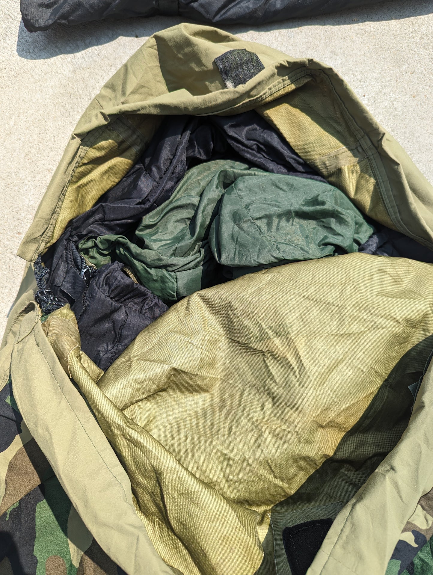 A top-down view looking into a large, open nylon carrying bag, showing the tightly packed components of the U.S. Military Modular Sleep System (MSS), including the black and olive green sleeping bags.