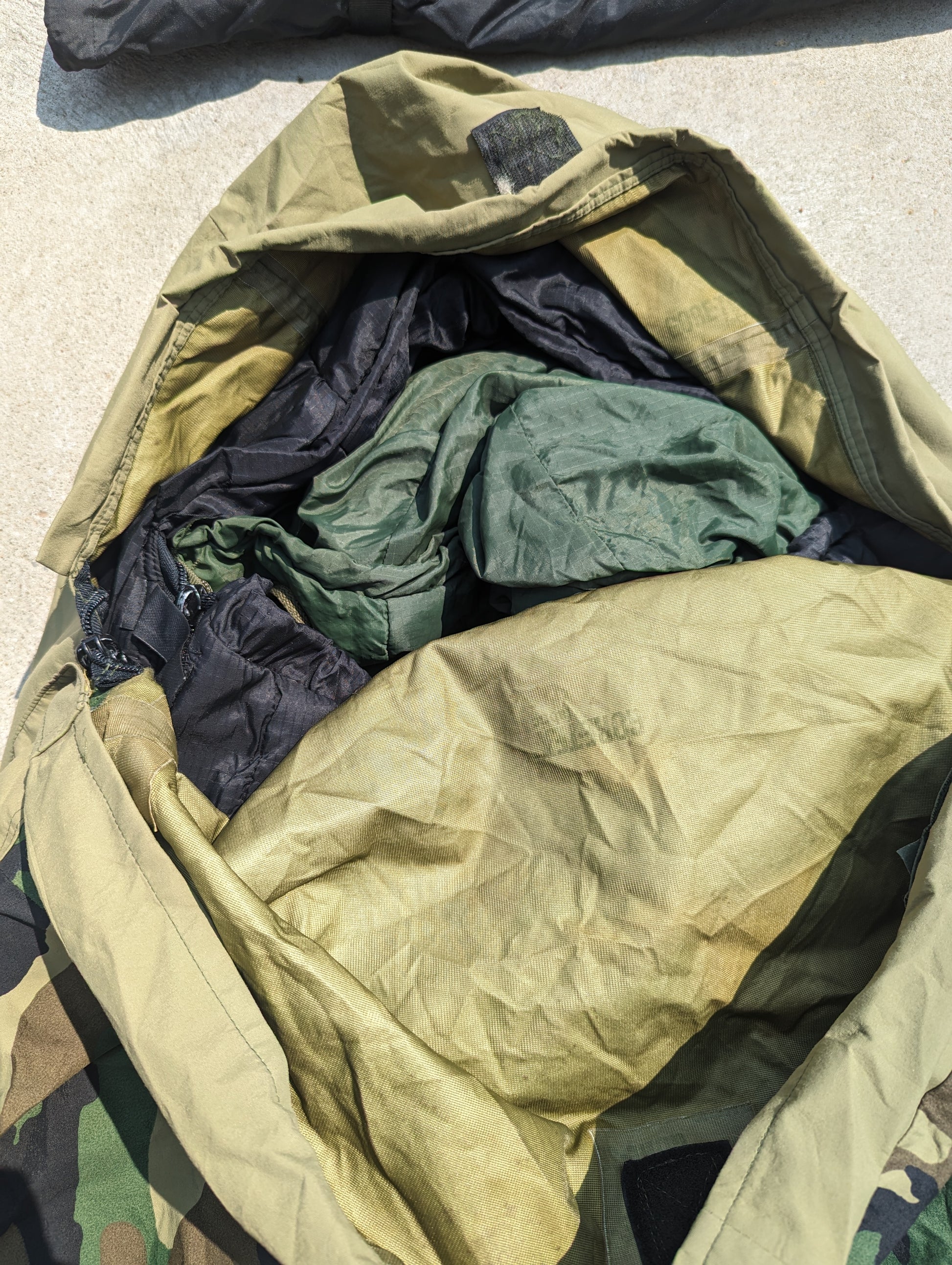 A top-down view looking into a large, open nylon carrying bag, showing the tightly packed components of the U.S. Military Modular Sleep System (MSS), including the black and olive green sleeping bags.