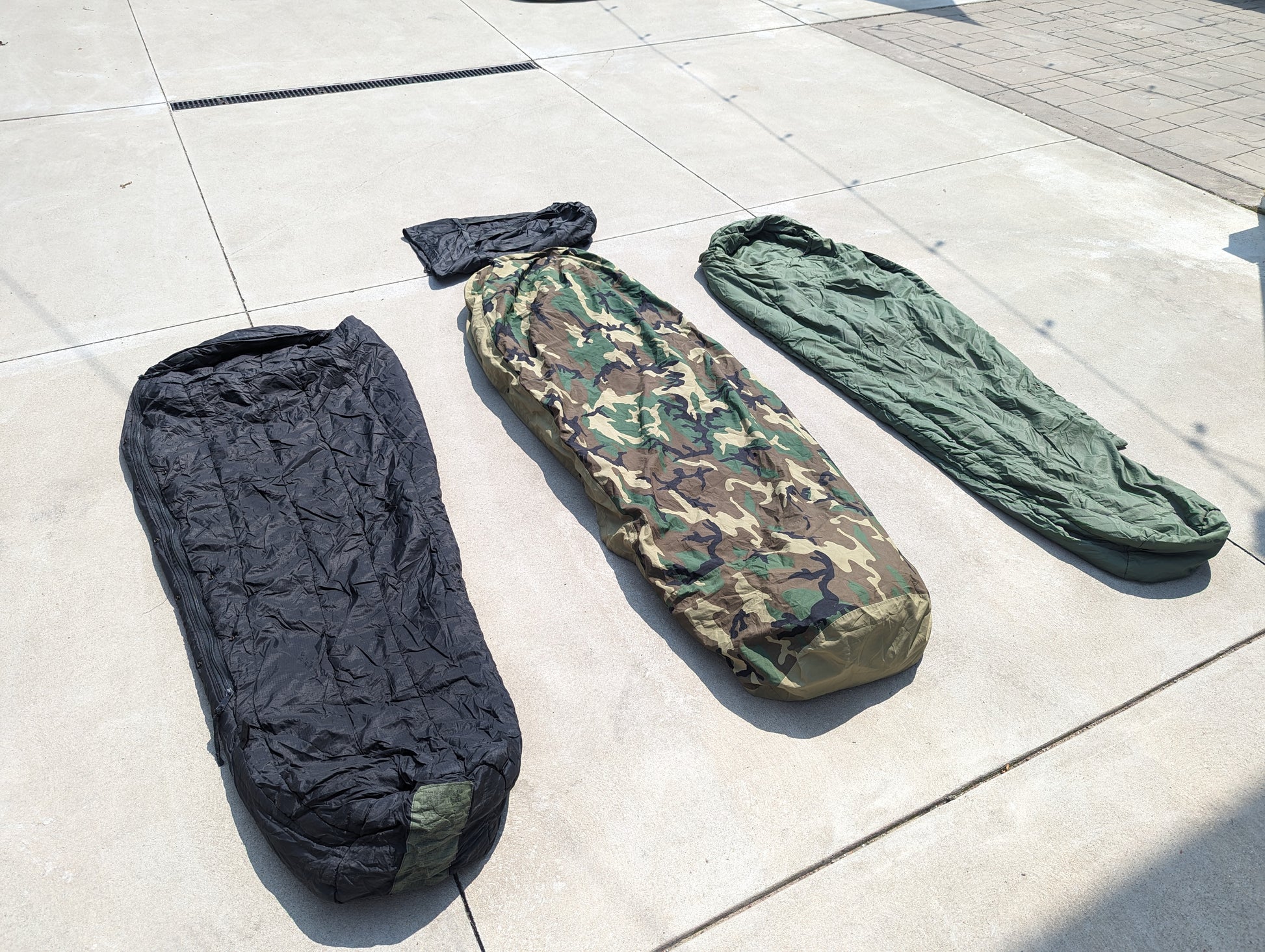 The three main layers of the U.S. Military Modular Sleep System (MSS) laid out on concrete, featuring the black Extreme Cold Weather bag, the Woodland camouflage component, and the solid olive green bag.