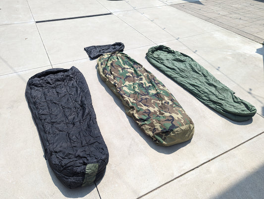 Three components of the U.S. Military Modular Sleep System (MSS) laid out on concrete, featuring the black sleeping bag, the Woodland camouflage cover, and the solid olive green bag.