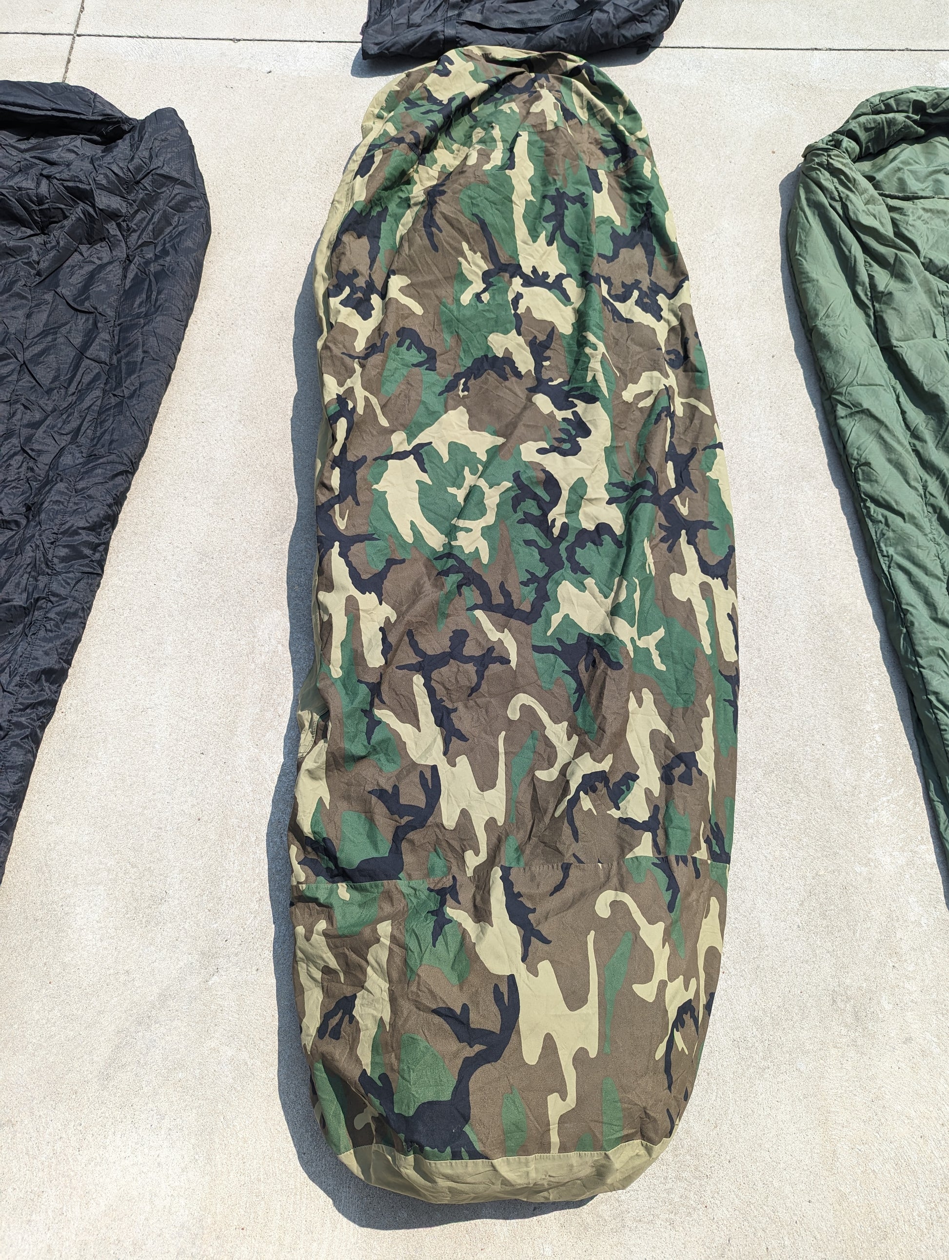 A full-length U.S. Military Modular Sleep System (MSS) component in Woodland camouflage, laid flat on a concrete surface, displaying the zippered, mummy-shaped cover.