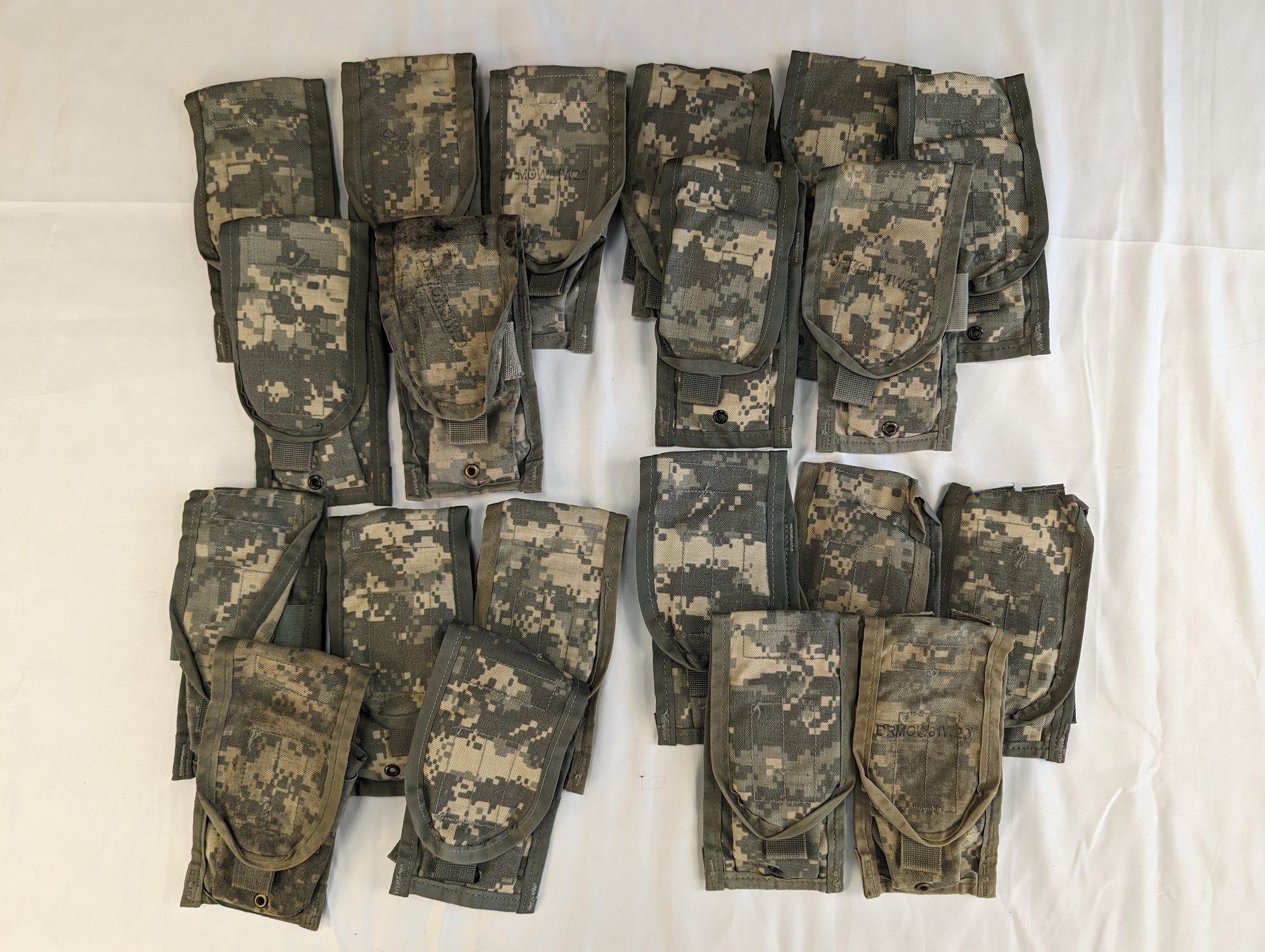 An overhead studio shot of approximately fifteen folded U.S. Military Surplus 2-Mag Pouches in ACU digital camouflage, laid out in an organized grid pattern on a white fabric background.