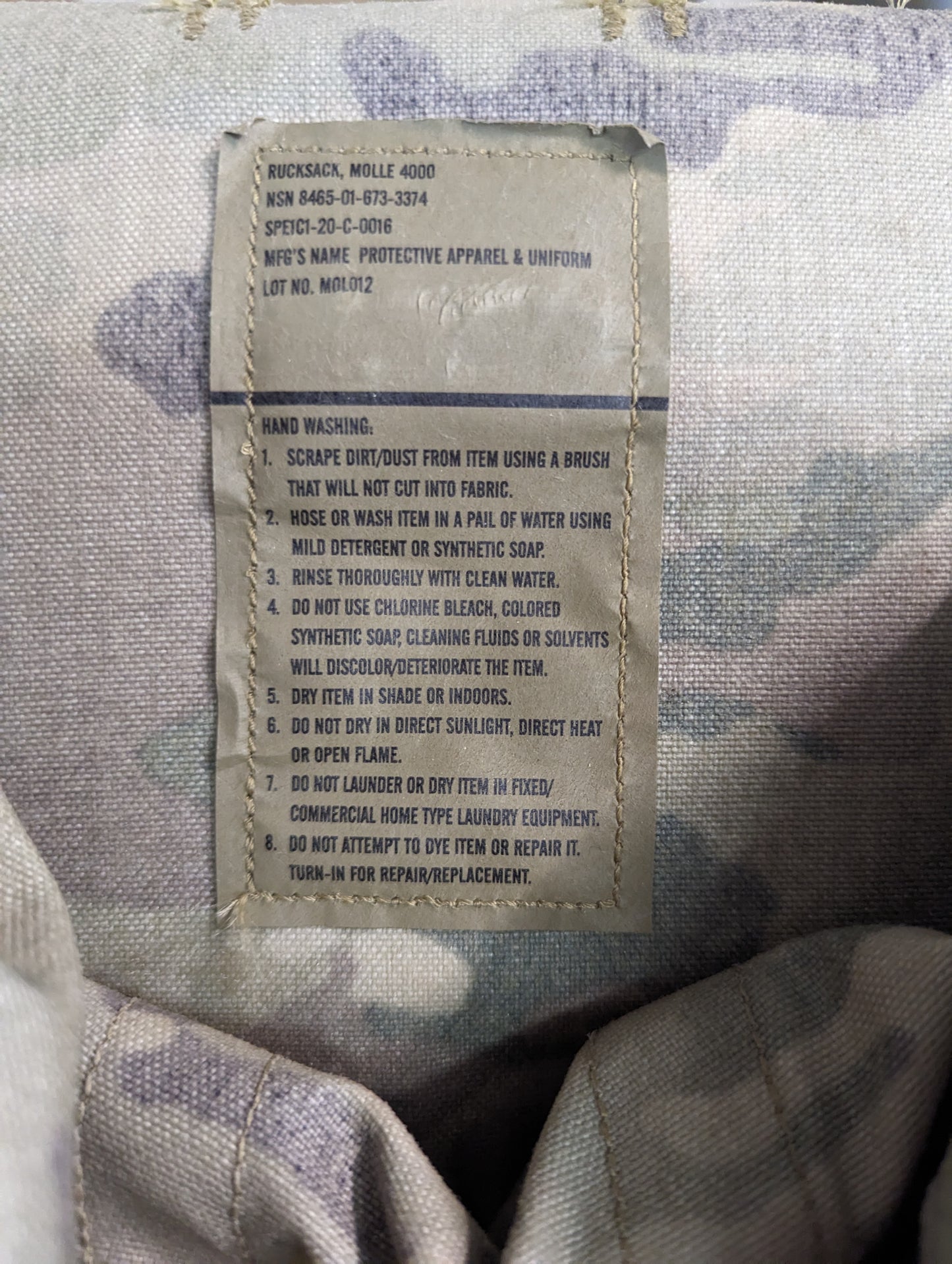A close-up photograph of the original manufacturer's care tag sewn into the OCP/Multicam MOLLE 4000 Rucksack, showing detailed hand washing and drying instructions.