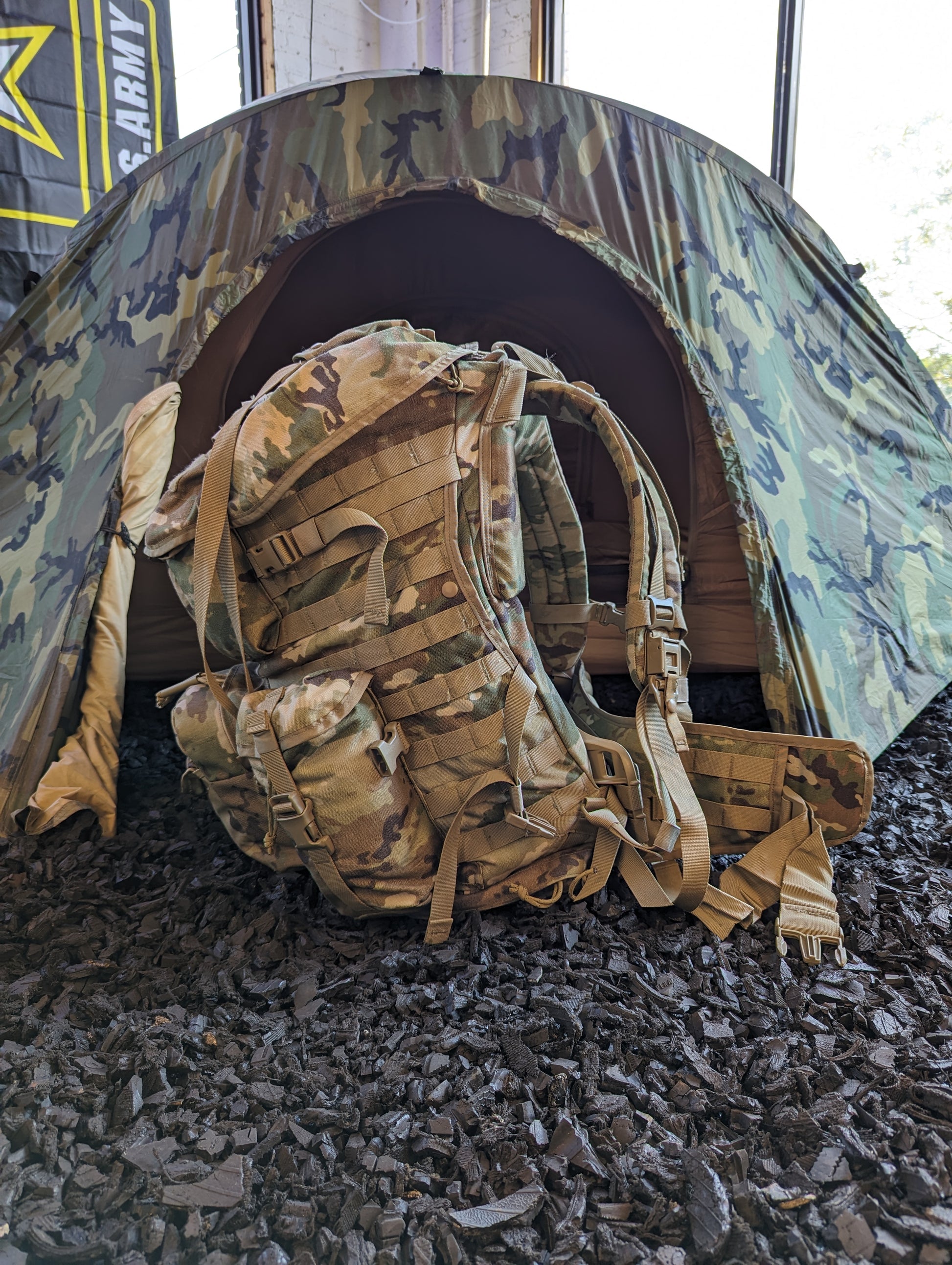 A dirty, field-used U.S. Military Surplus MOLLE 4000 Rucksack in OCP/Multicam camouflage lying on its side next to a partially set up U.S. Army camouflage tent.