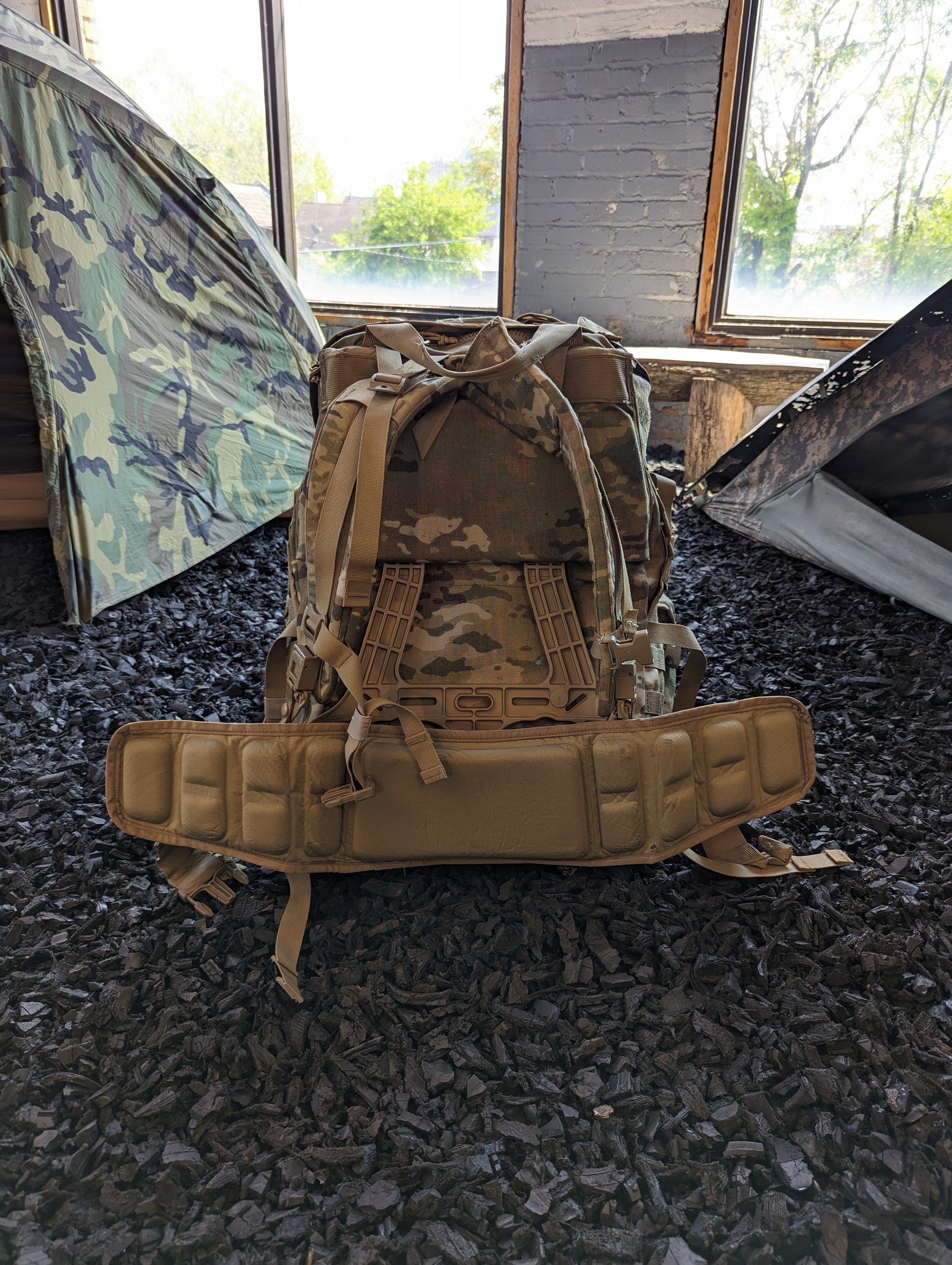 A large U.S. Military Surplus MOLLE 4000 Rucksack in OCP/Multicam camouflage, heavily covered in mud and dirt, resting on the ground in an abandoned indoor setting, highlighting its rugged, used condition.