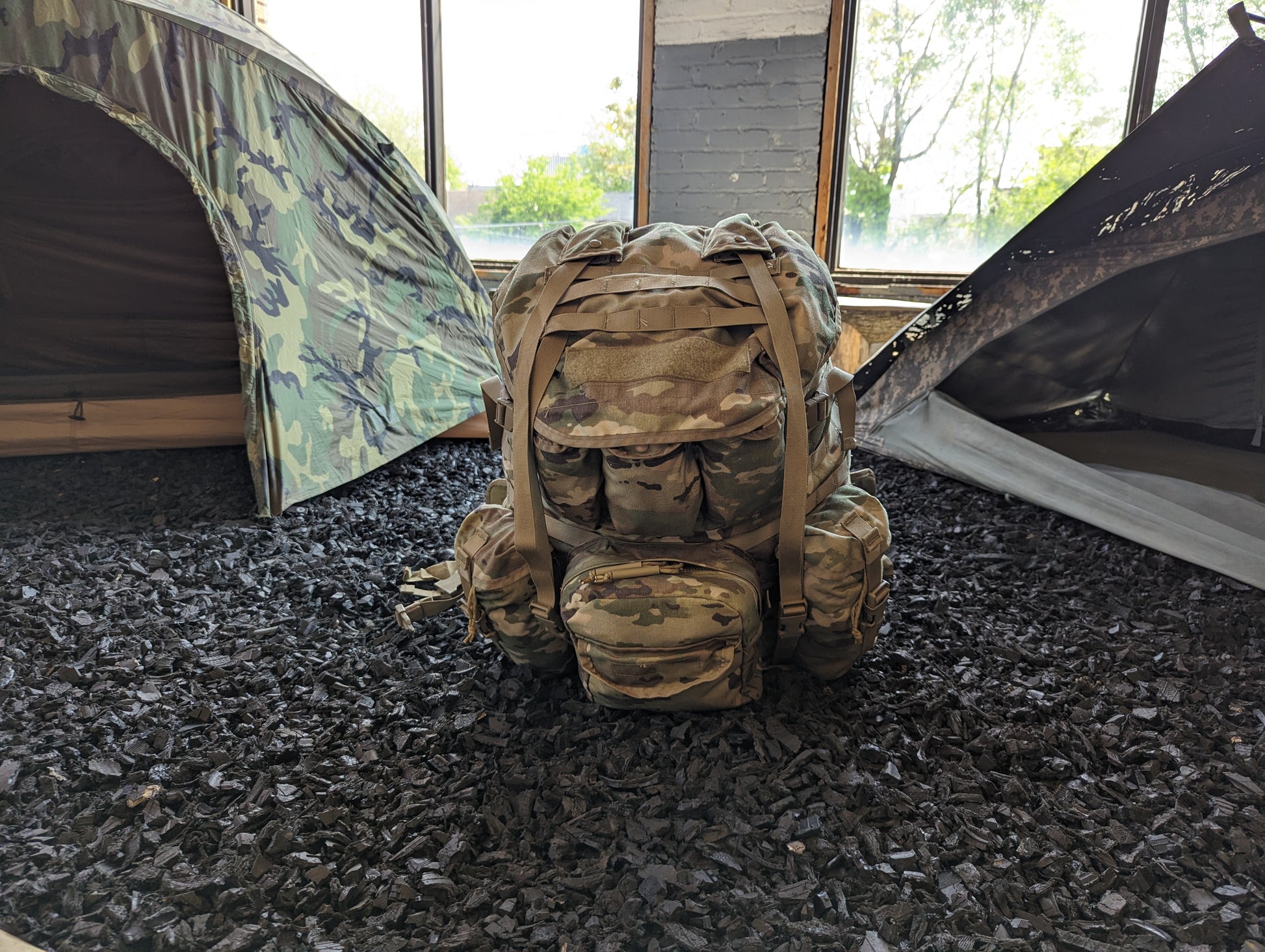 A MOLLE 4000 Large Field Pack in OCP/Multicam camouflage standing on a floor of dark mulch, set in an interior space with a partially disassembled camo tent in the background.