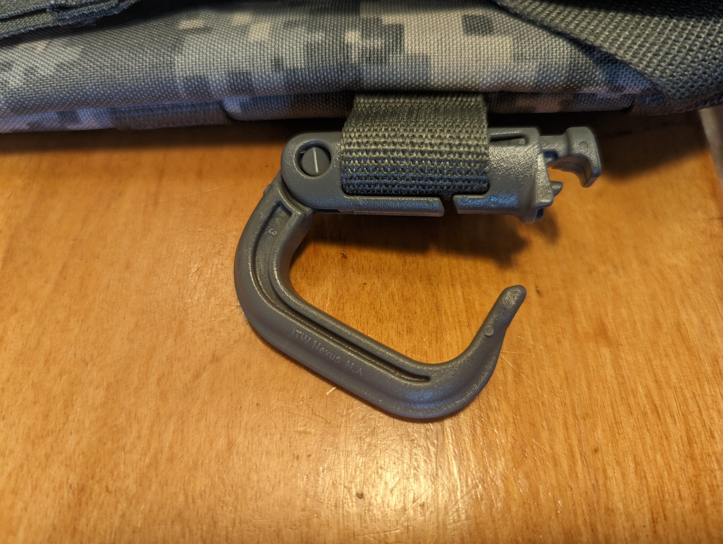 A close-up detail shot of a gray, plastic D-ring carabiner (Grimloc-style) secured to an olive green webbing strap on an ACU camouflage pack, showcasing the twist-to-lock mechanism.