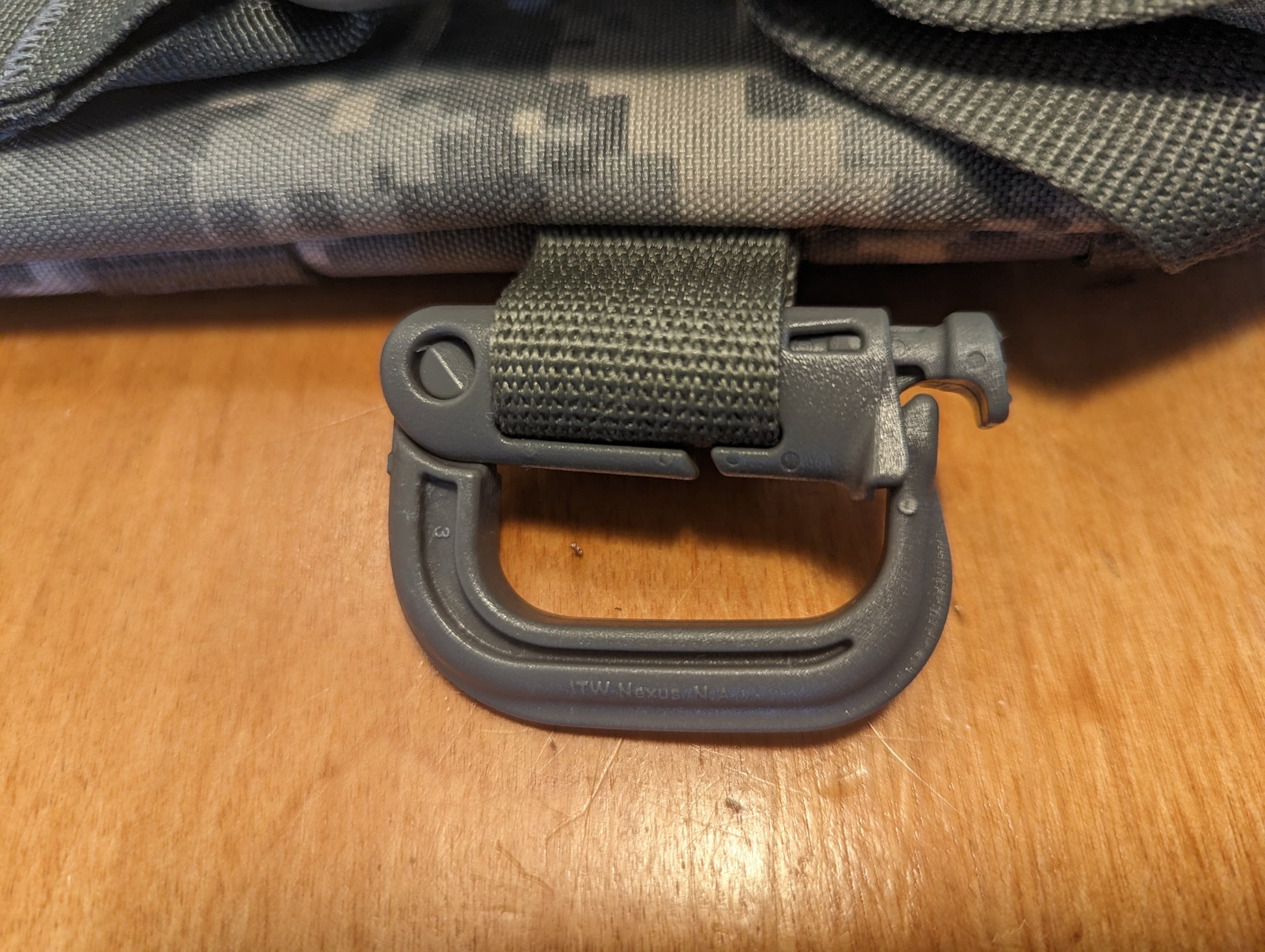 A close-up detail shot of a gray, plastic D-ring carabiner (Grimloc-style) secured to an olive green webbing strap on an ACU camouflage pack, showcasing the twist-to-lock mechanism.