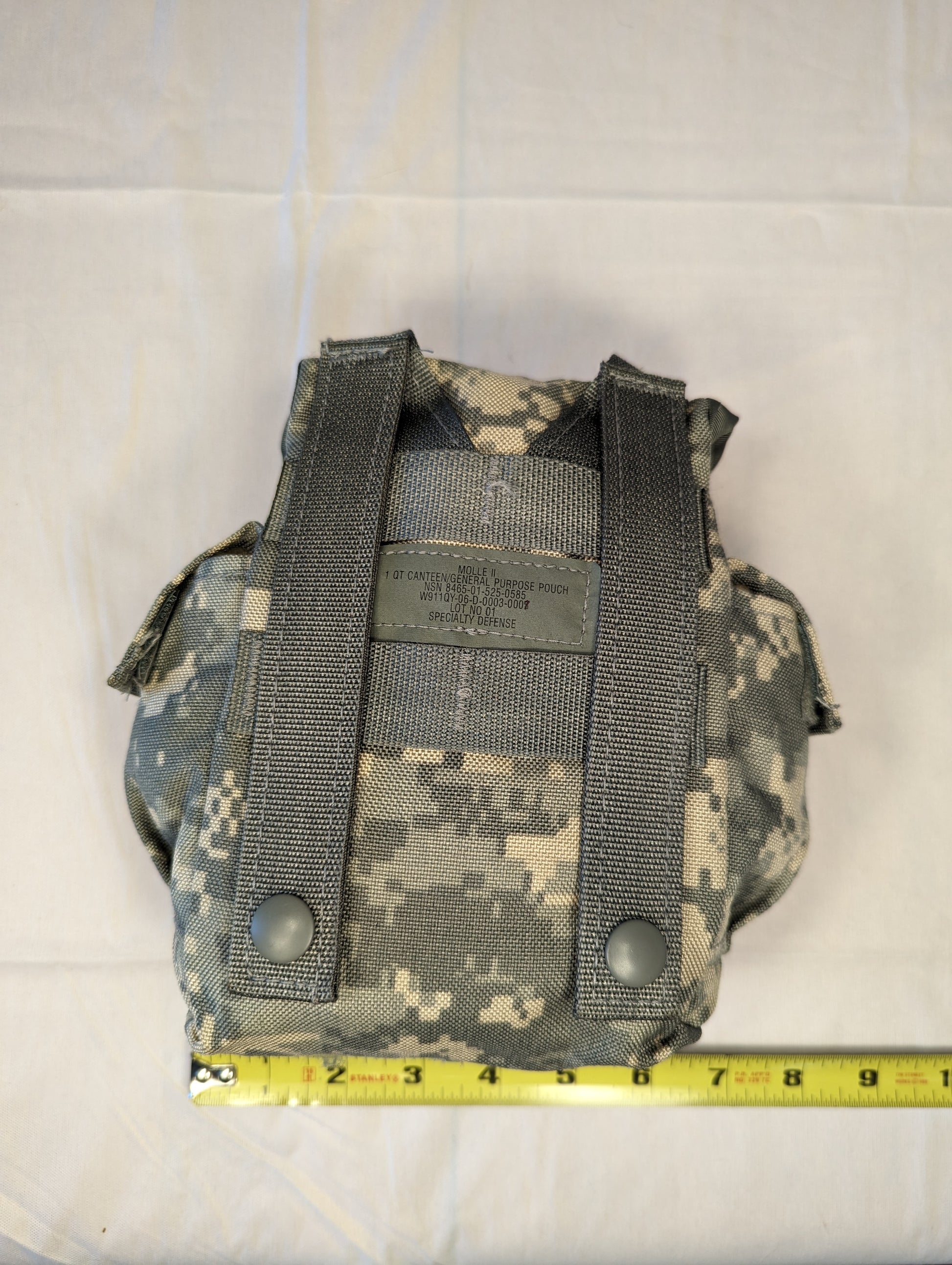 A back view of a U.S. Military Surplus MOLLE II 1-Quart Canteen Pouch in ACU digital camouflage, displaying the vertical MOLLE attachment straps and a yellow tape measure indicating the width.