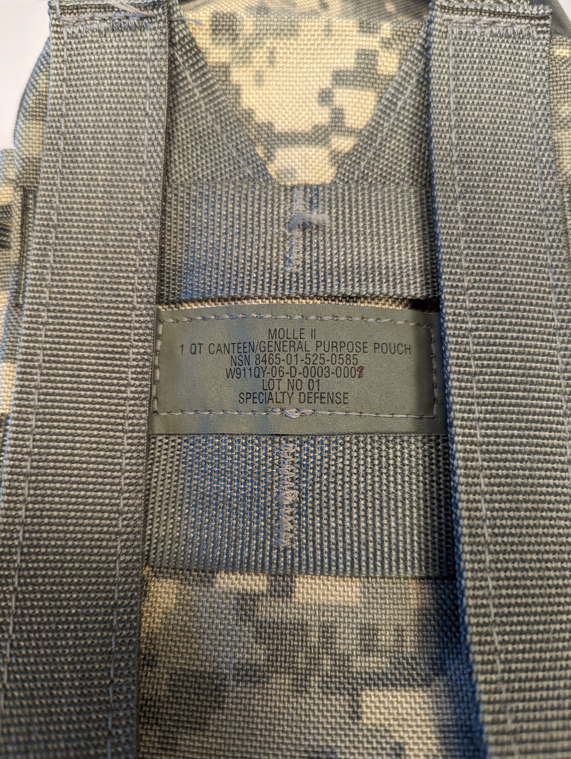 A close-up photograph of the official U.S. Military issue tag sewn onto the MOLLE webbing of an ACU camouflage Canteen/General Purpose Pouch, clearly displaying the product name, NSN, and manufacturer 'SPECIALTY DEFENSE'.