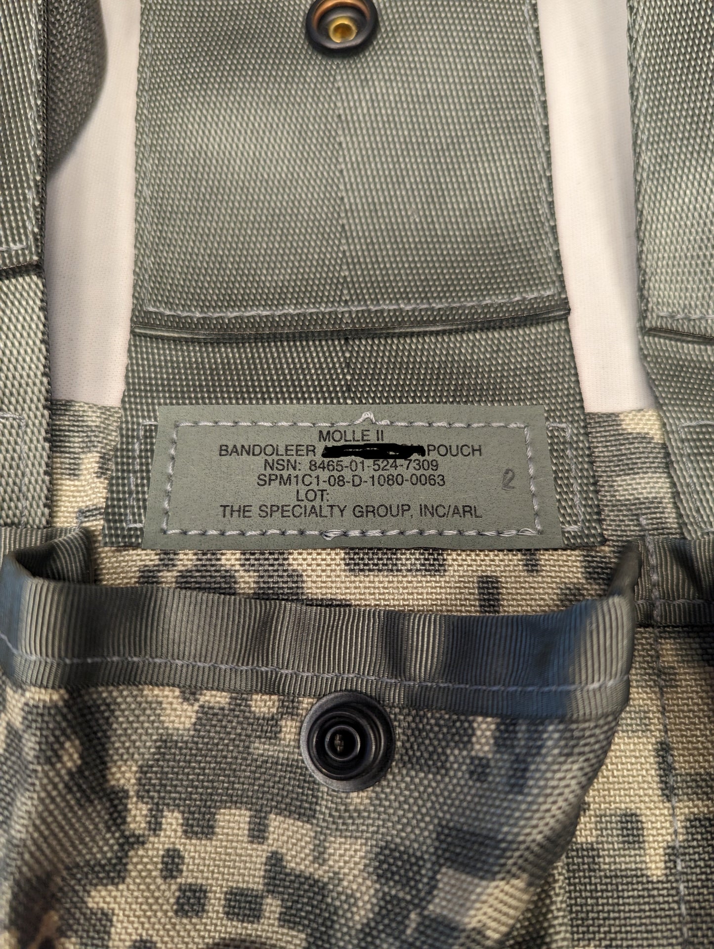 A close-up photograph of the official U.S. Military issue tag sewn into the ACU digital camouflage Bandoleer Pouch, displaying 'MOLLE II BANDOLEER AMMUNITION POUCH', NSN 8465-01-524-7309, and manufacturer 'THE SPECIALTY GROUP, INC.'.