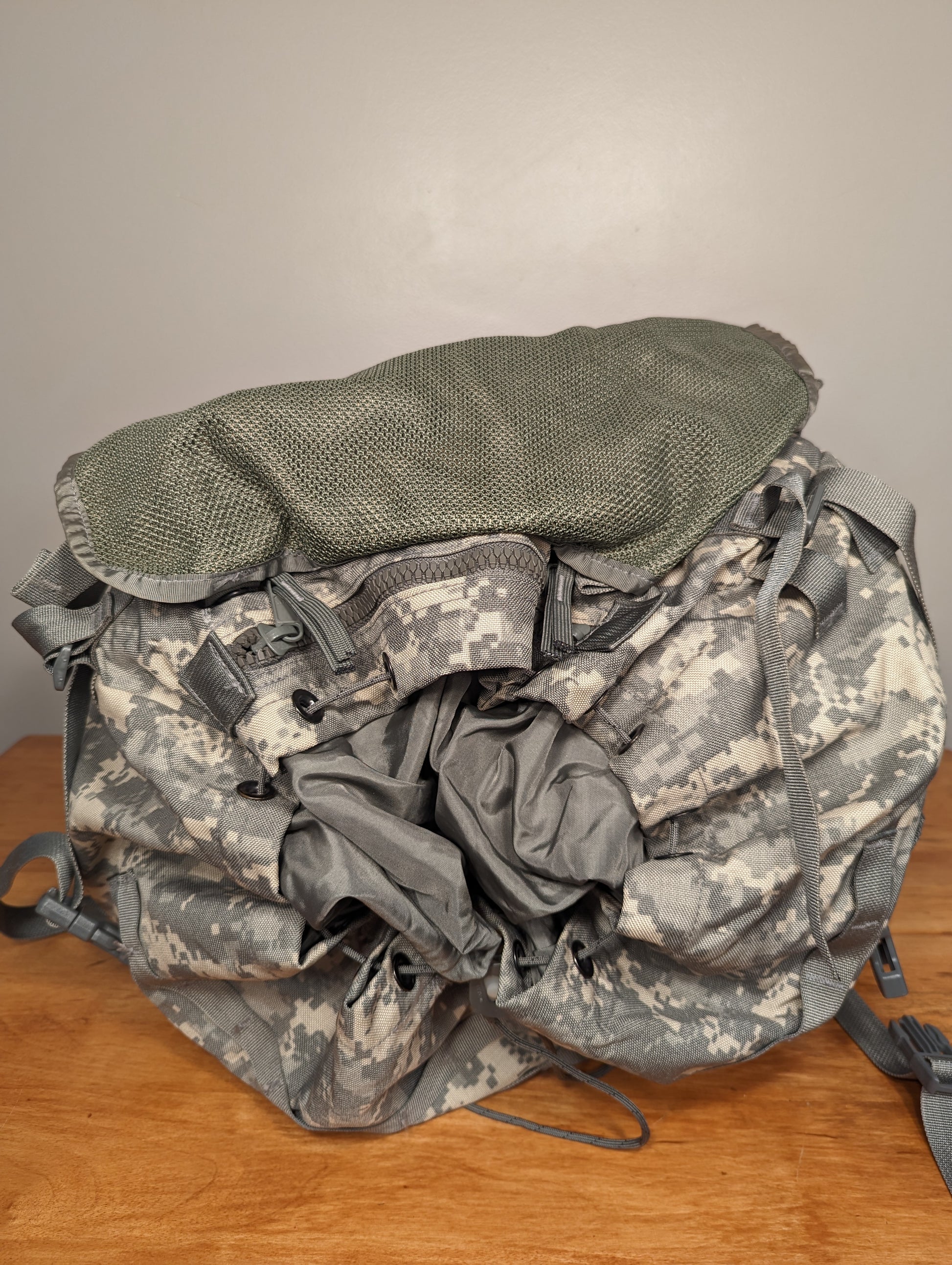 A front view of a U.S. Military Surplus MOLLE II Large Rucksack in ACU digital camouflage, resting on a wooden surface with the top flap flipped back, revealing the textured foliage green lining and drawstring storm collar.