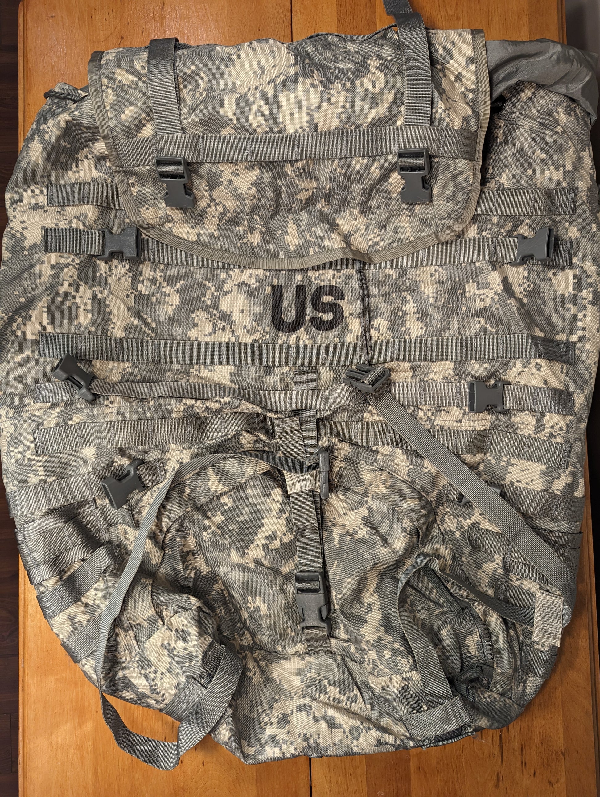 A front view of a U.S. Military Surplus MOLLE II Large Rucksack in ACU digital camouflage, lying flat on a wooden surface, featuring a large black 'US' stencil on the top flap, extensive horizontal webbing, and multiple compression straps.