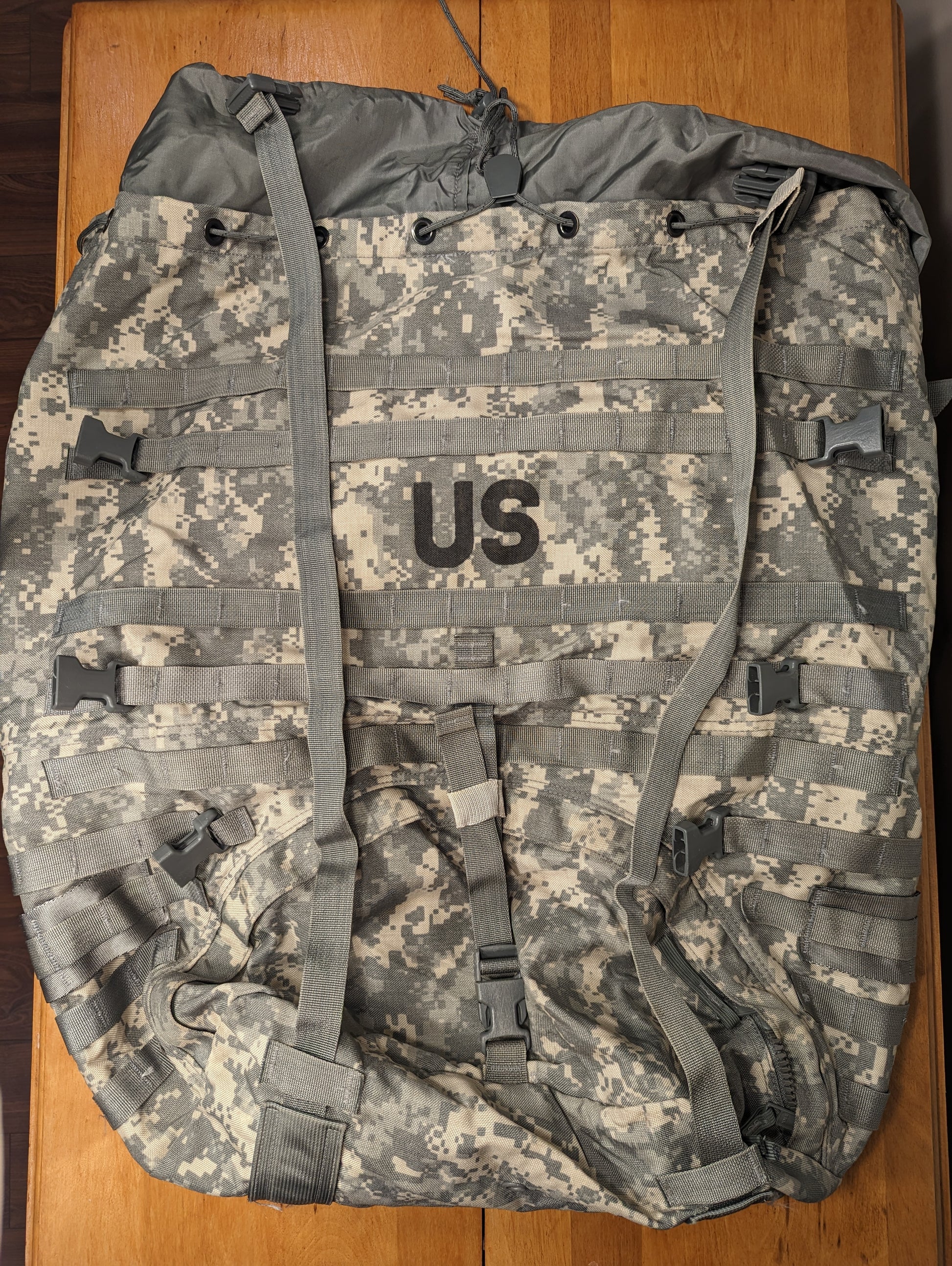 A front view of a U.S. Military Surplus MOLLE II Large Rucksack in ACU digital camouflage, lying flat on a wooden surface, featuring a large black 'US' stencil on the top flap, extensive horizontal webbing, and gray quick-release buckles.