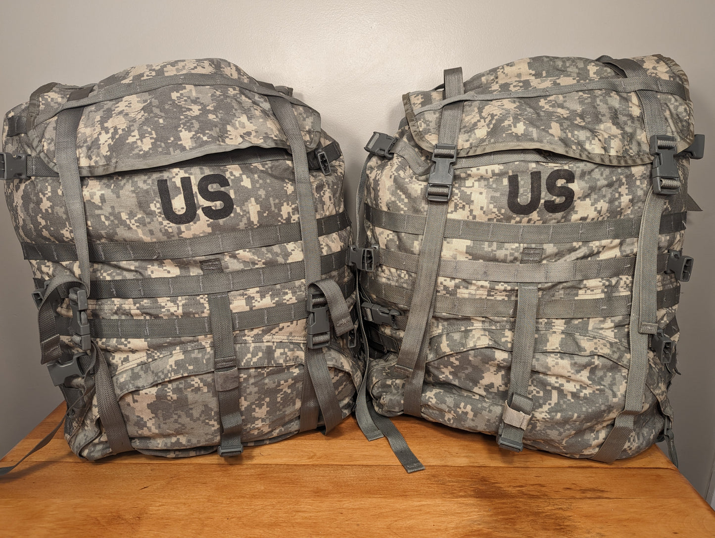 Two U.S. Military Surplus MOLLE II Large Rucksacks in ACU digital camouflage, standing upright side-by-side on a wooden surface, featuring large black 'US' stencils on the top flaps and extensive webbing.
