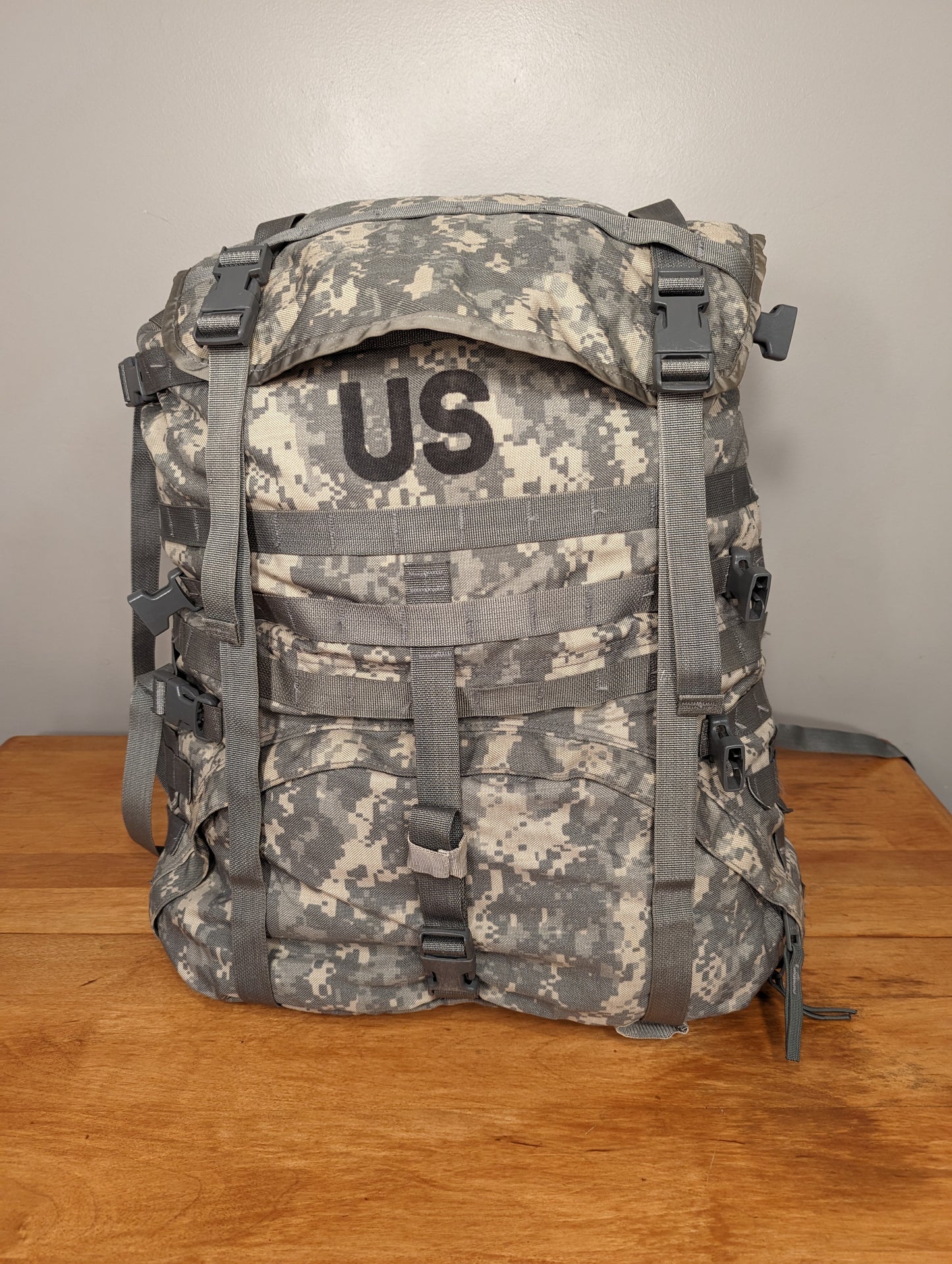 A front view of a U.S. Military Surplus MOLLE II Large Rucksack in ACU/UCP digital camouflage, standing upright on a wooden surface, featuring extensive horizontal MOLLE webbing, gray quick-release buckles, and a large black stenciled 'US' mark on the top flap.