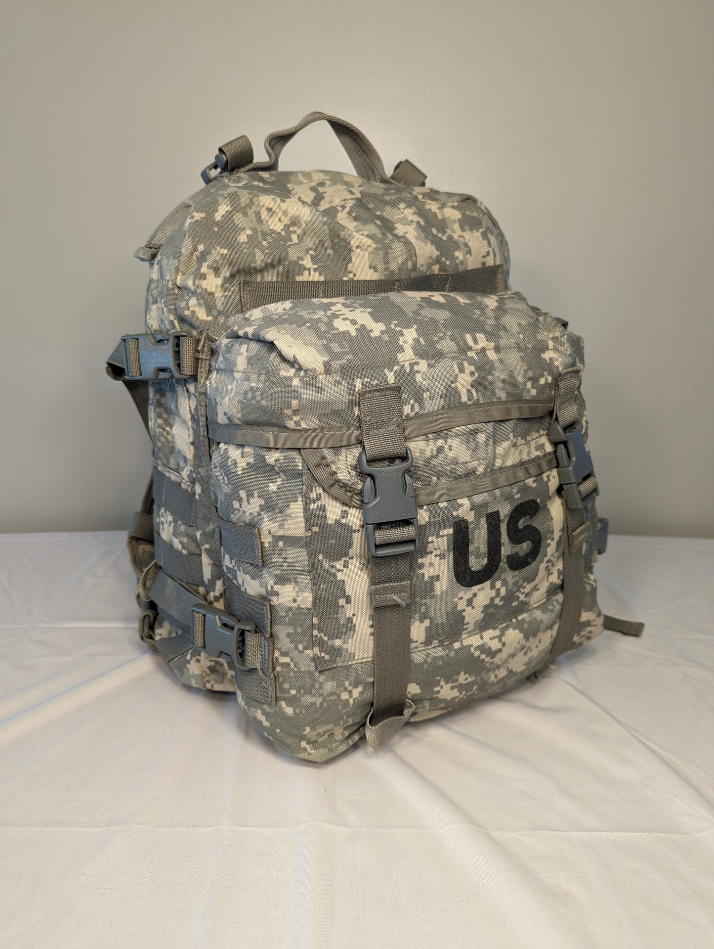 A large U.S. Military Surplus MOLLE II Rucksack in ACU digital camouflage, standing upright, featuring a large black stenciled 'US' mark on the front, extensive MOLLE webbing, and heavy-duty buckles.