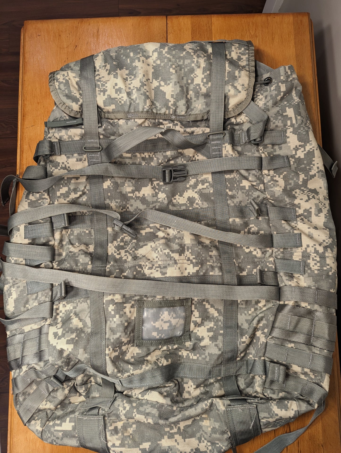 A large U.S. Military Surplus MOLLE II Rucksack in ACU digital camouflage, laid flat on a wooden table, showcasing the full structure, extensive horizontal MOLLE webbing, and numerous compression straps.
