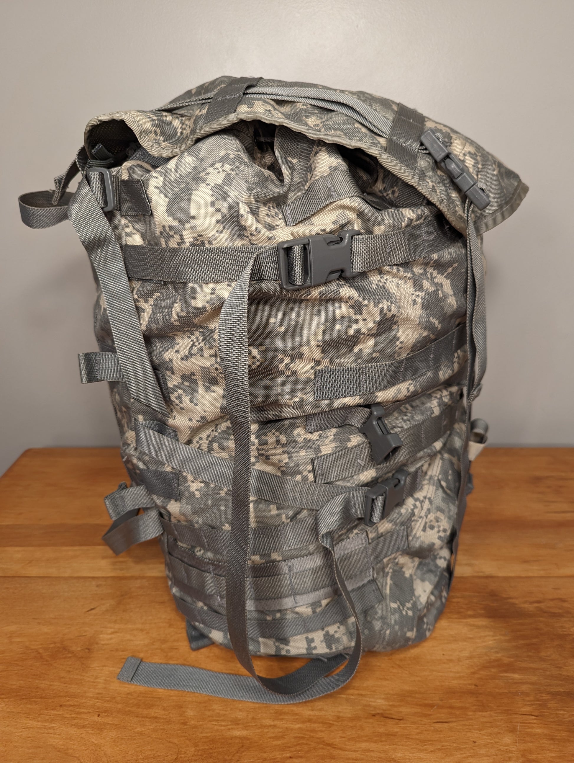 A U.S. Military Surplus MOLLE II Large Rucksack in ACU digital camouflage, standing upright on a wooden surface, featuring extensive webbing loops, gray compression straps, and a large main compartment.