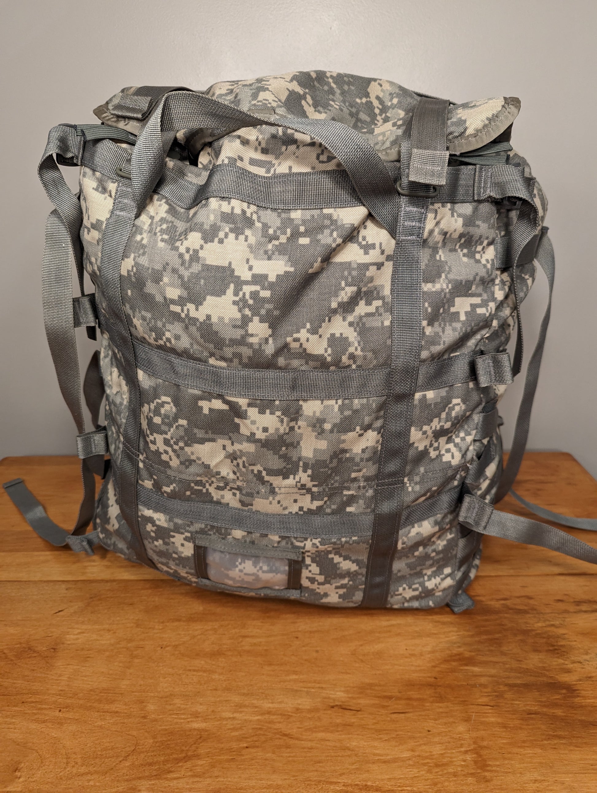 A front view of a U.S. Military Surplus MOLLE II Large Rucksack in ACU/UCP digital camouflage, standing upright on a wooden surface, featuring extensive horizontal webbing, gray compression straps, and a clear identification window at the bottom.