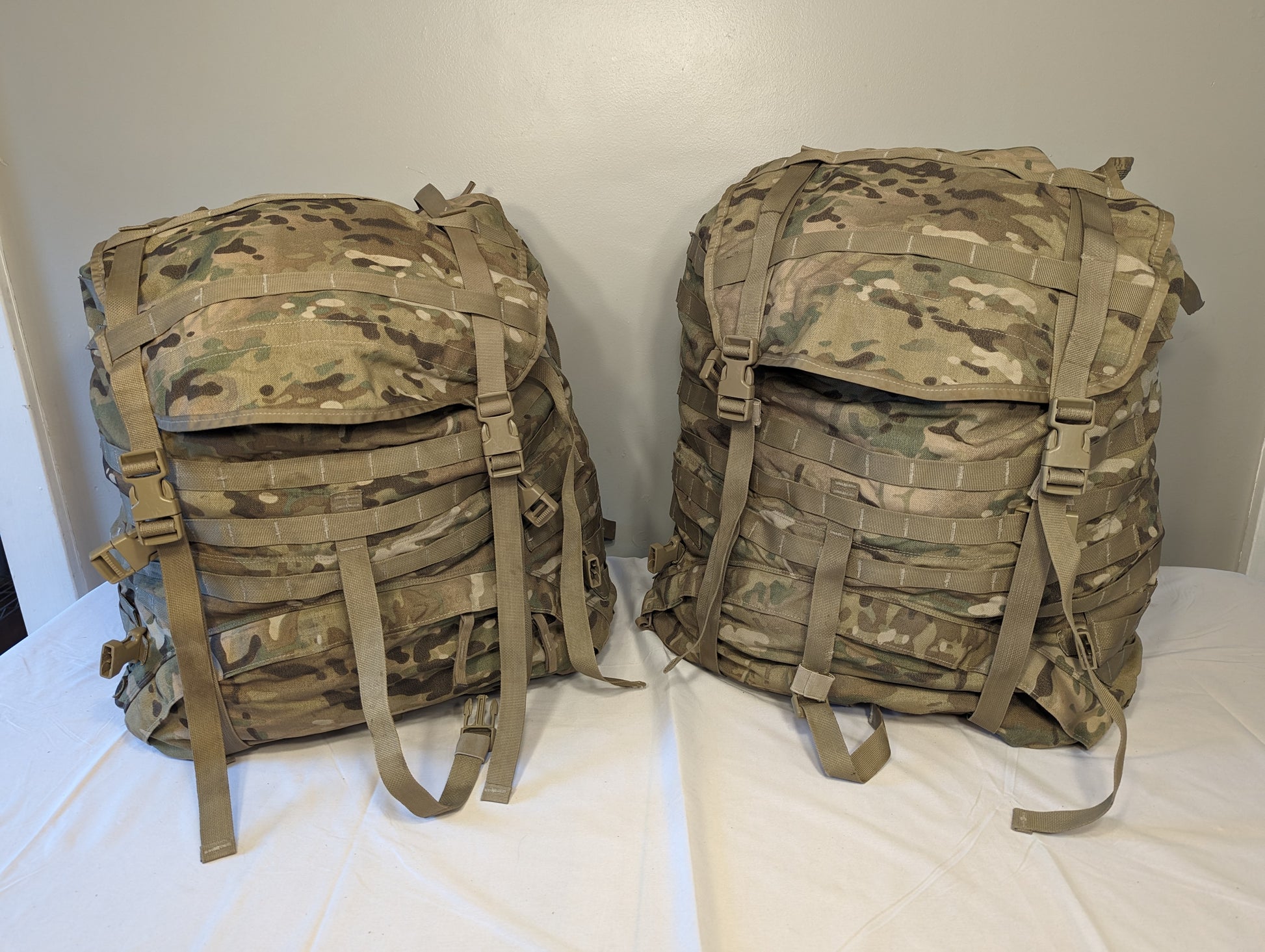 Two U.S. Military Surplus MOLLE II Large Rucksacks in OCP/Multicam camouflage, standing upright side-by-side on a white surface, featuring large flap closures, extensive webbing, and compression straps.