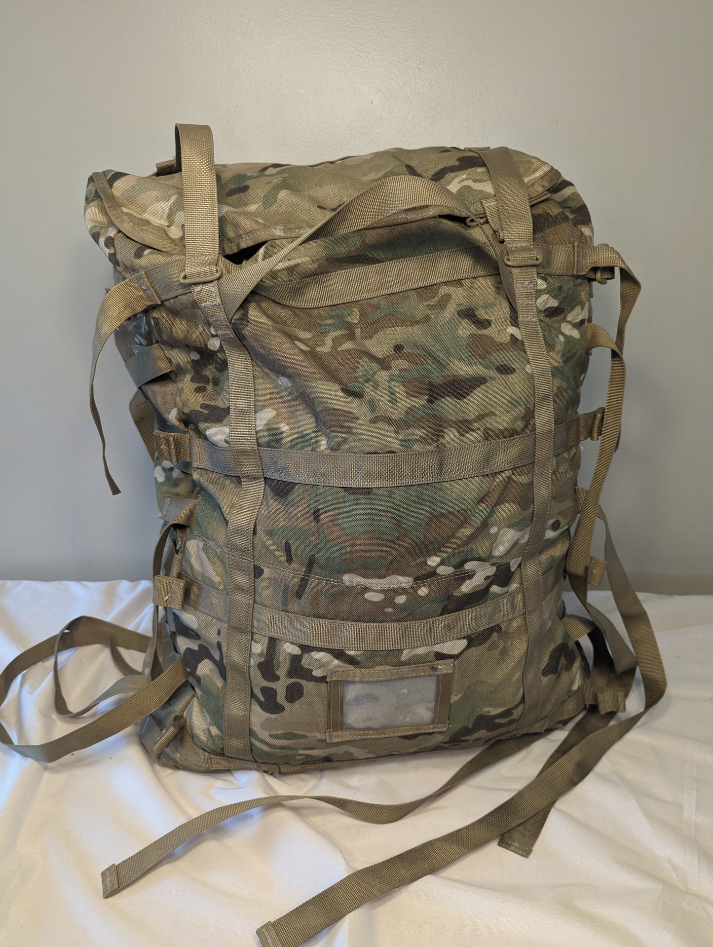 A rear view of a U.S. Military Surplus MOLLE II Large Rucksack in OCP/Multicam camouflage, standing upright, featuring extensive horizontal webbing, vertical straps, and a clear identification window.