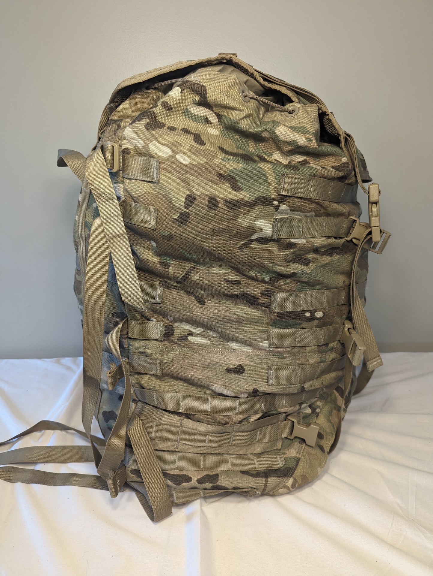 A side view of a U.S. Military Surplus MOLLE II Large Rucksack in OCP/Multicam camouflage, standing upright on a white surface, featuring extensive horizontal webbing, vertical compression straps, and a large top flap.