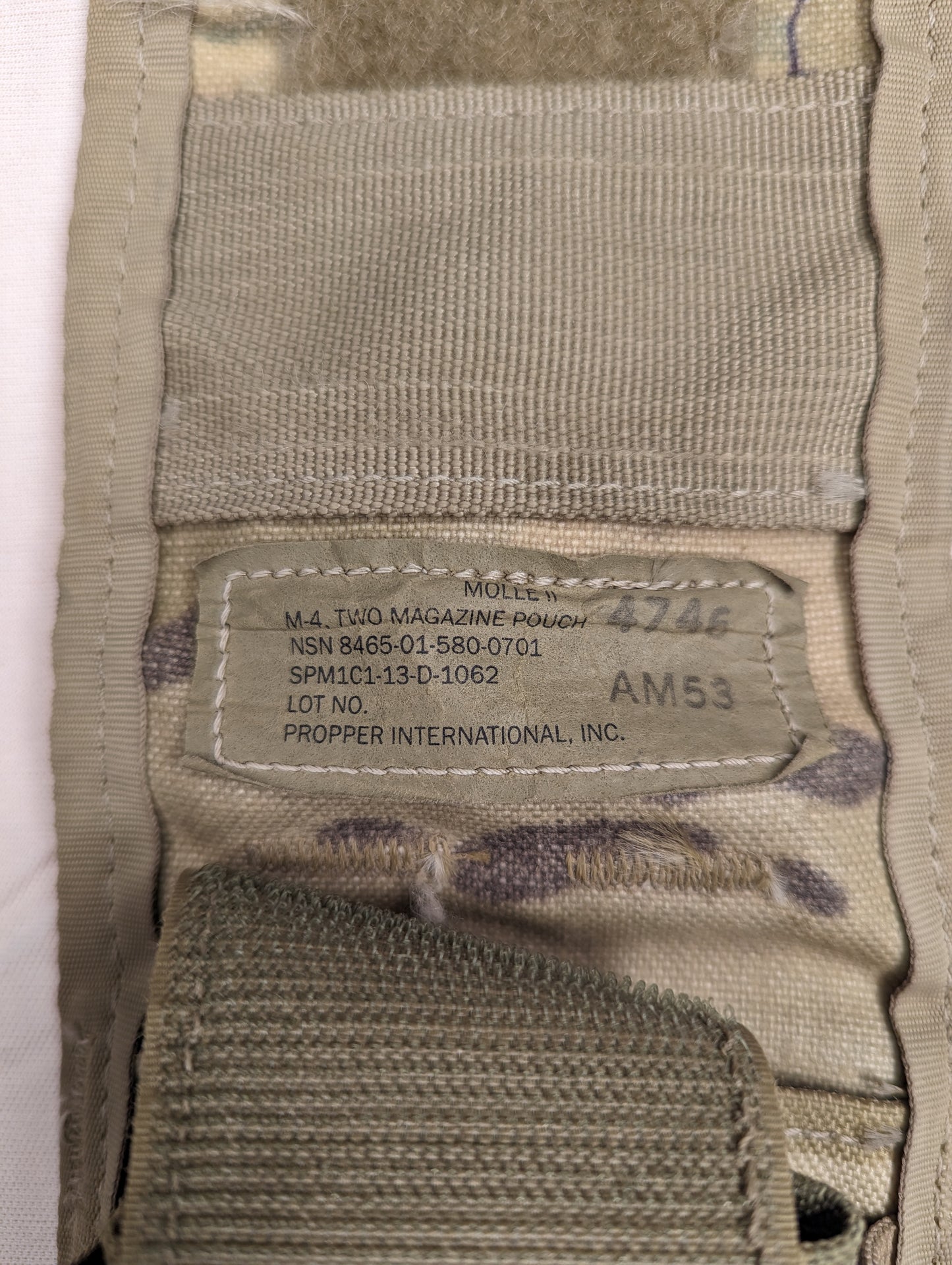 A close-up photograph of the official U.S. Military issue tag sewn into the OCP/Multicam Double Magazine Pouch, displaying 'MOLLE II M-4 TWO MAGAZINE POUCH', NSN 8465-01-580-0701, and manufacturer 'PROPPER INTERNATIONAL, INC.'.