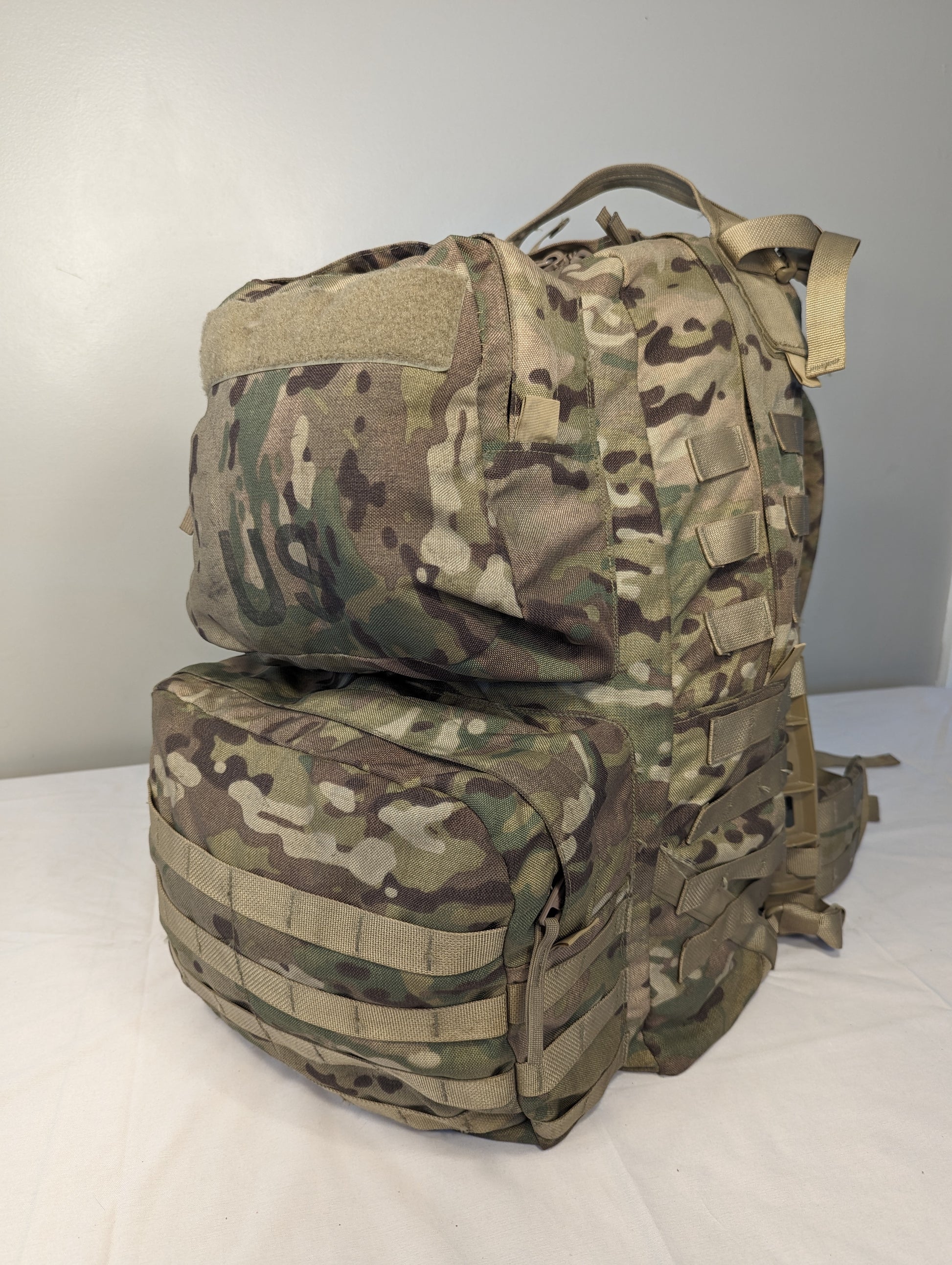 A side-front view of a U.S. Military Surplus Medium Field Pack Rucksack in OCP/Multicam camouflage, featuring a large upper compartment, a separate lower compartment, and extensive horizontal MOLLE webbing.