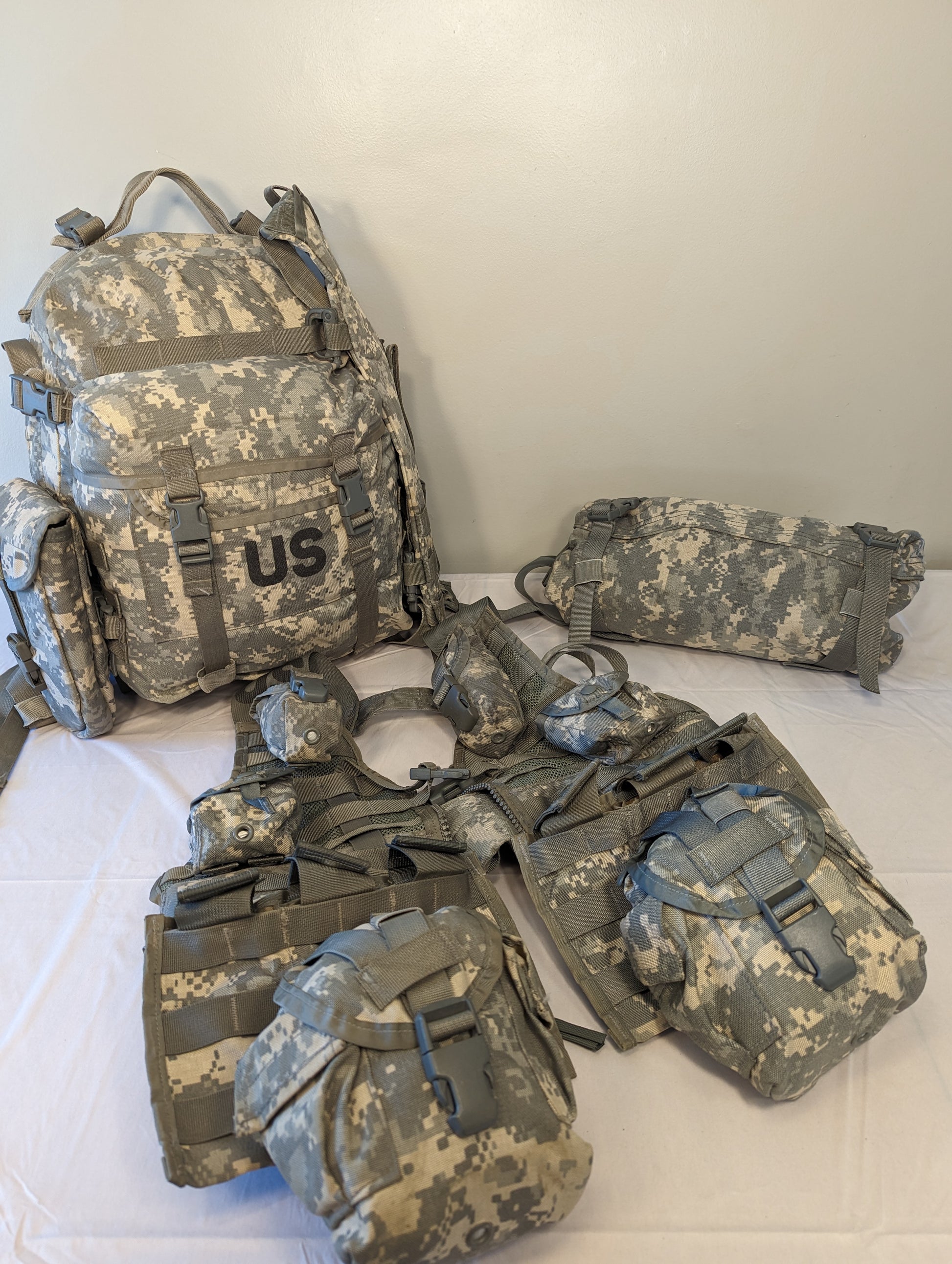 An overhead flat lay photograph of a complete U.S. Military MOLLE II Rifleman Set in ACU digital camouflage. The kit includes a large rucksack with a prominent 'US' stencil, a tactical vest, and numerous attached and detached accessory pouches.