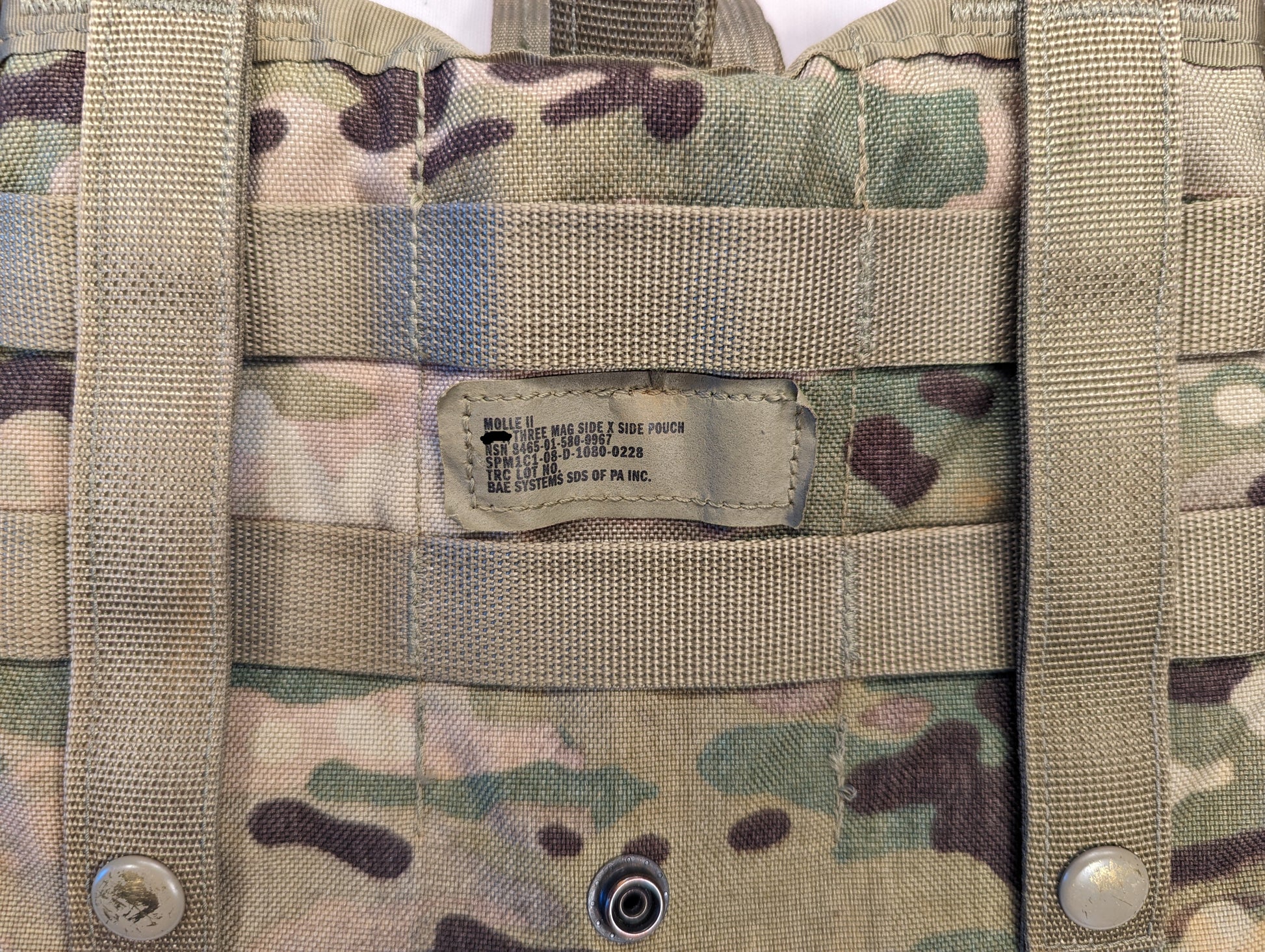 A close-up photograph of a U.S. Military OCP/Multicam MOLLE pouch, displaying the manufacturer's issue tag with NSN and company name 'BAL SYSTEMS LTD/KAY PT, INC.' underneath the horizontal MOLLE webbing.