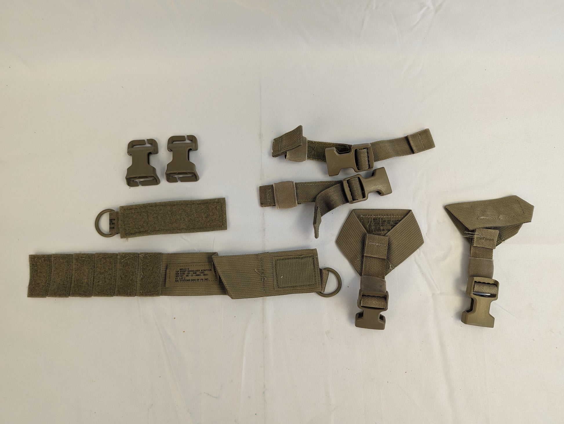 An overhead view of a set of detachable U.S. Military Surplus tactical webbing components, featuring olive drab nylon straps, plastic quick-release buckles, and D-rings, laid out on a light background.