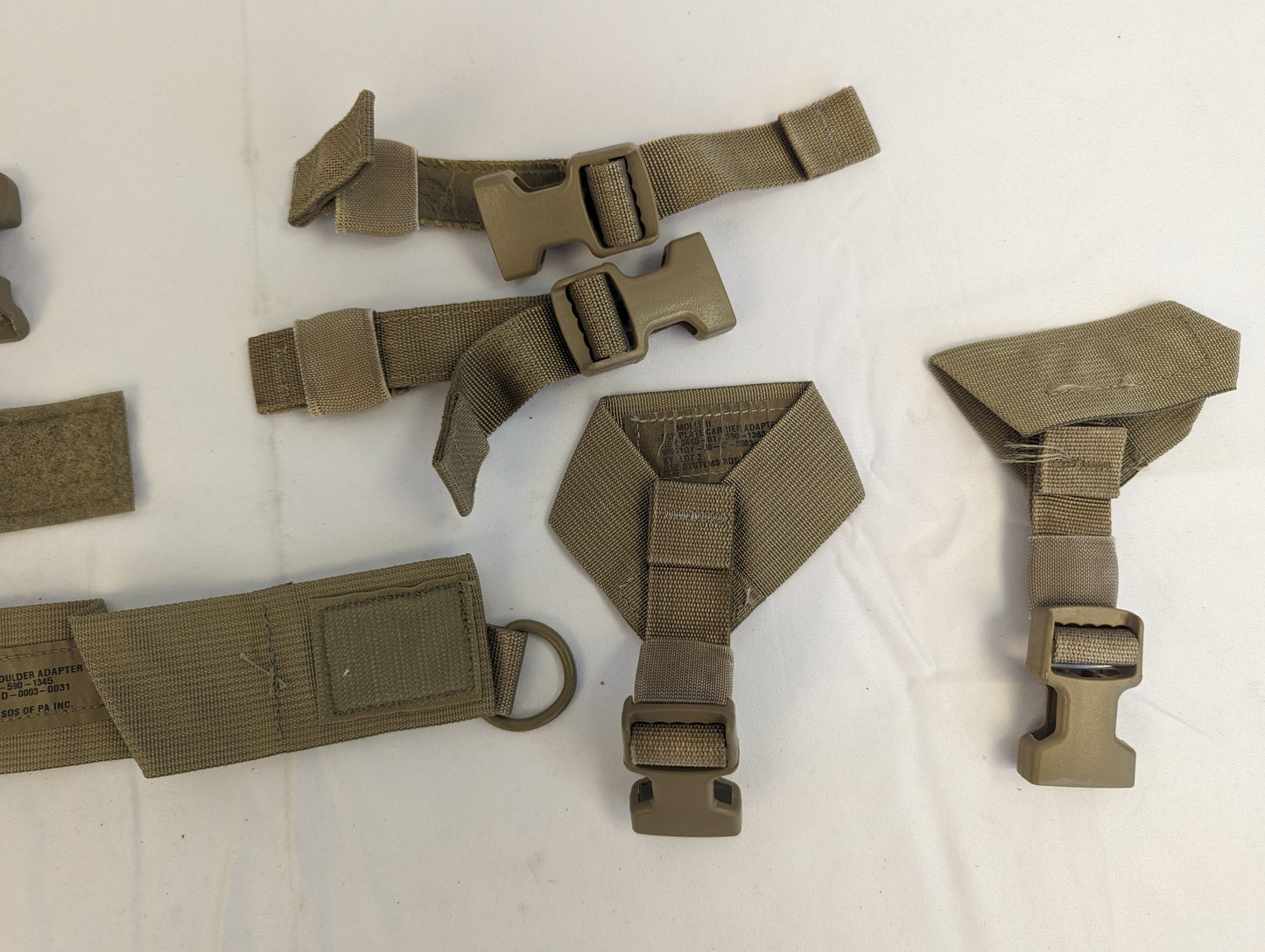 An overhead view of U.S. Military Surplus Coyote Brown MOLLE webbing and harness components, including quick-release buckles, adjustment straps, a large metal D-ring, and a specialized U-shaped adapter.