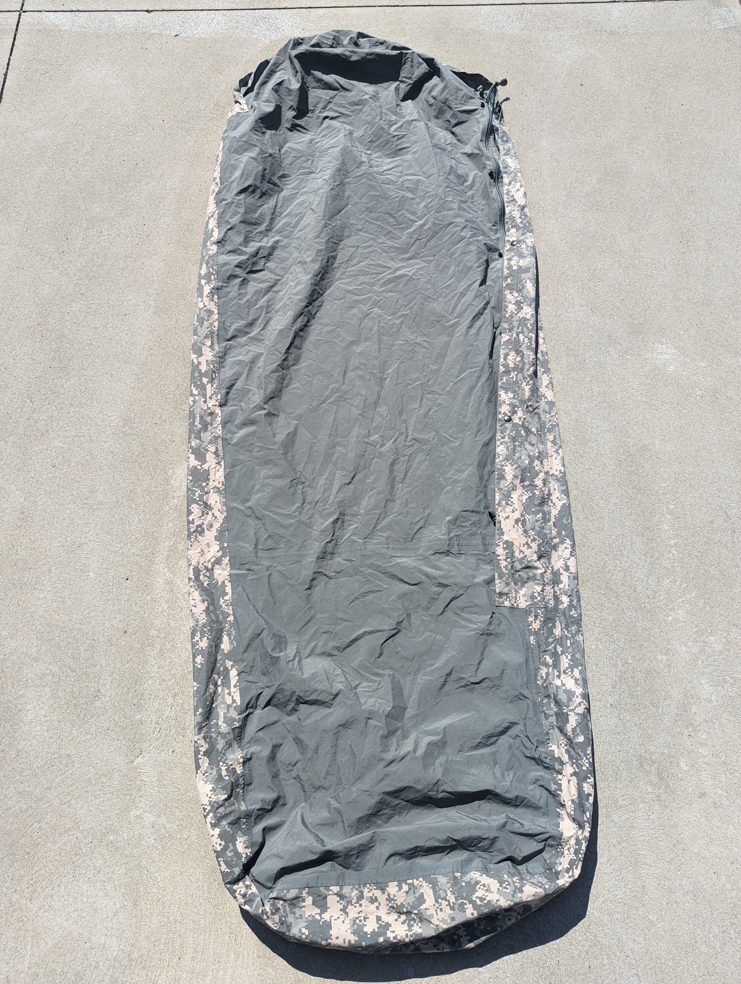 A full-length view of a U.S. Military Surplus Modular Sleep System Bivy Cover in ACU digital camouflage, zipped closed and laid flat on a gray concrete surface, displaying the mummy shape and wrinkled waterproof fabric.