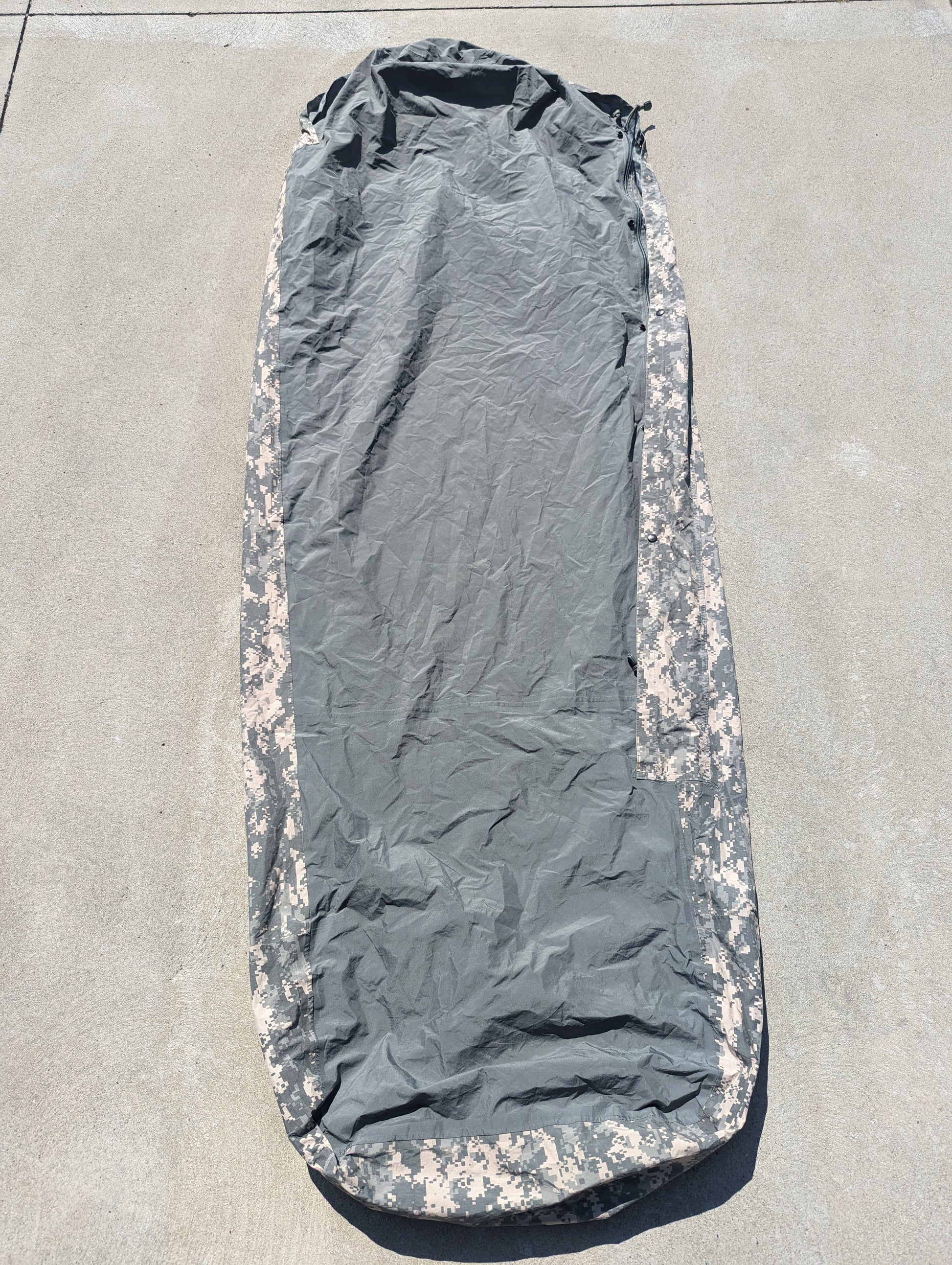 A full-length view of a U.S. Military Surplus Modular Sleep System Bivy Cover in ACU digital camouflage, zipped closed and laid flat on a gray concrete surface, displaying the mummy shape and wrinkled waterproof fabric.