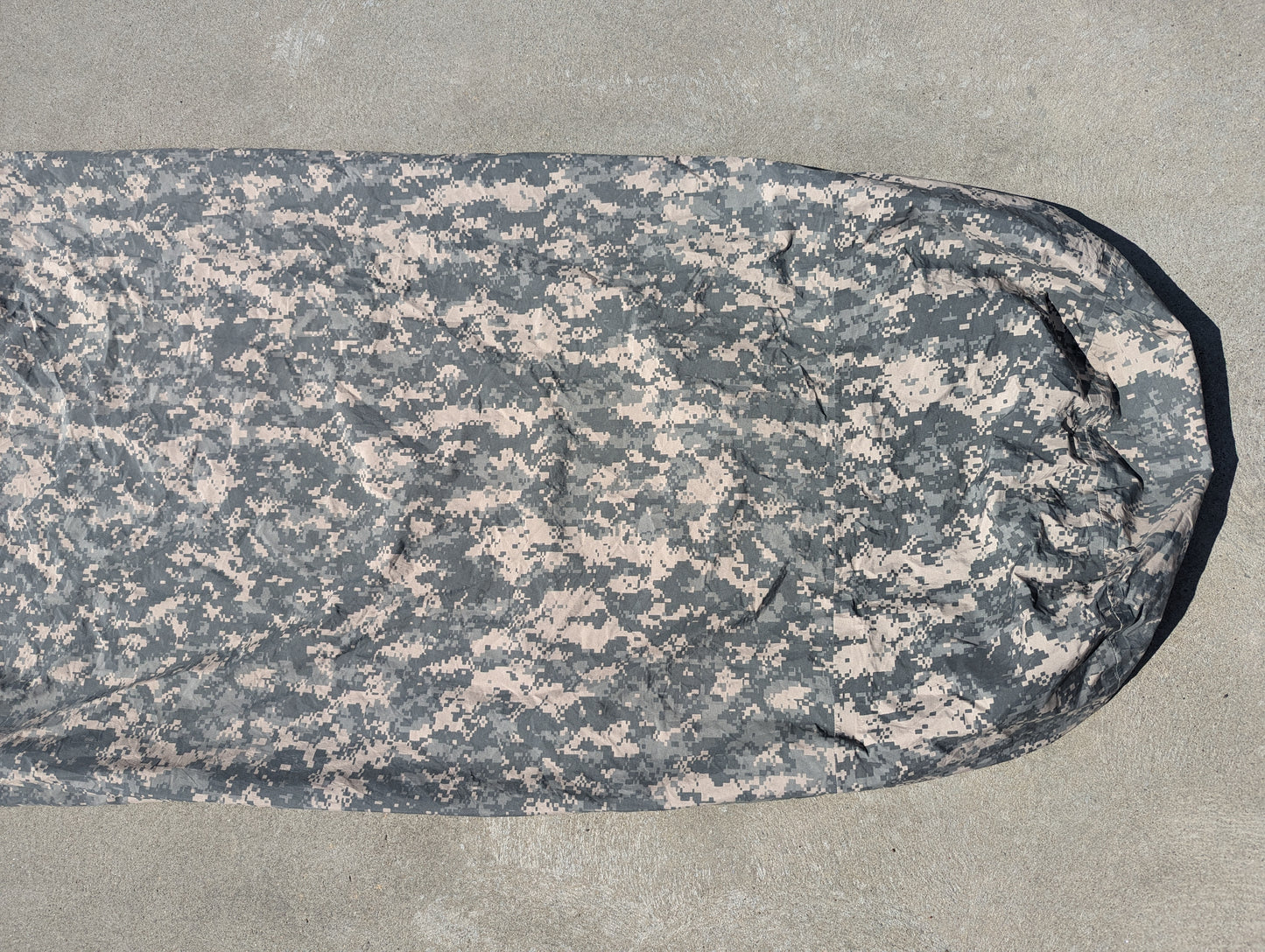 A full-length U.S. Military Surplus Modular Sleep System Bivy Cover in ACU digital camouflage, zipped closed and laid flat on a gray concrete background.