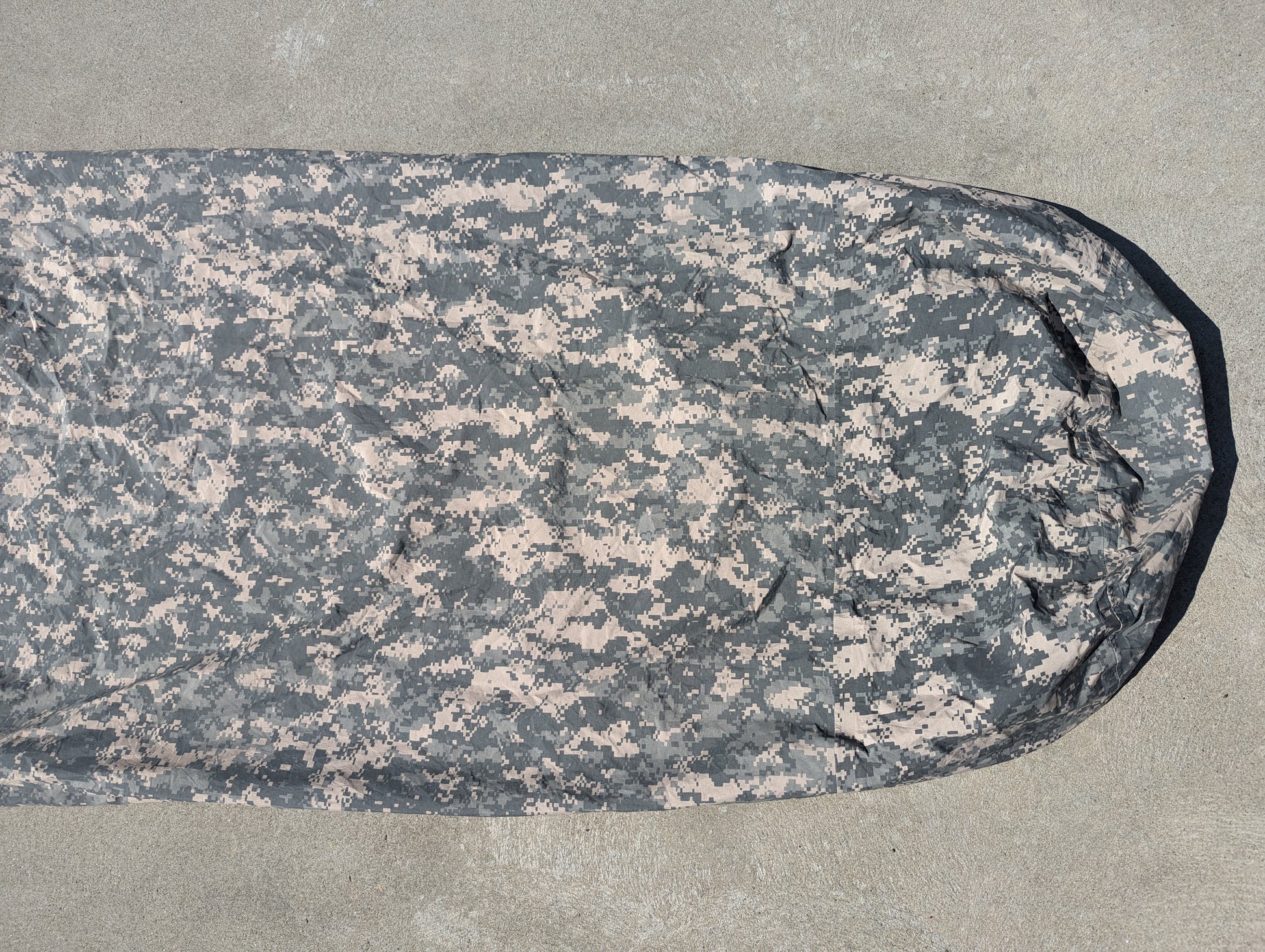 A full-length U.S. Military Surplus Modular Sleep System Bivy Cover in ACU digital camouflage, zipped closed and laid flat on a gray concrete background.