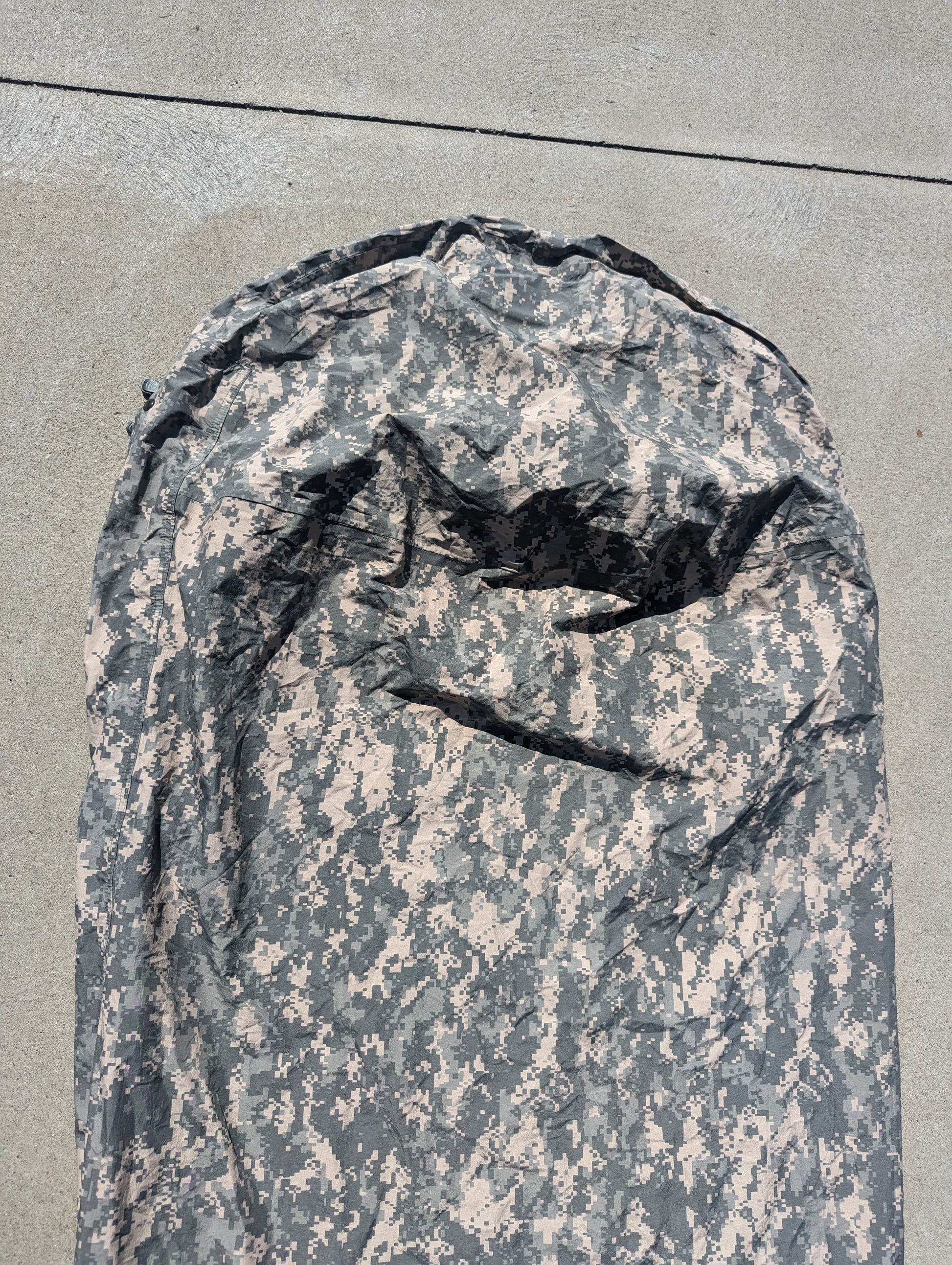 A full-length view of a U.S. Military Surplus Modular Sleep System Bivy Cover in ACU digital camouflage, zipped closed and laid flat on a gray concrete surface, displaying the size and camo pattern.