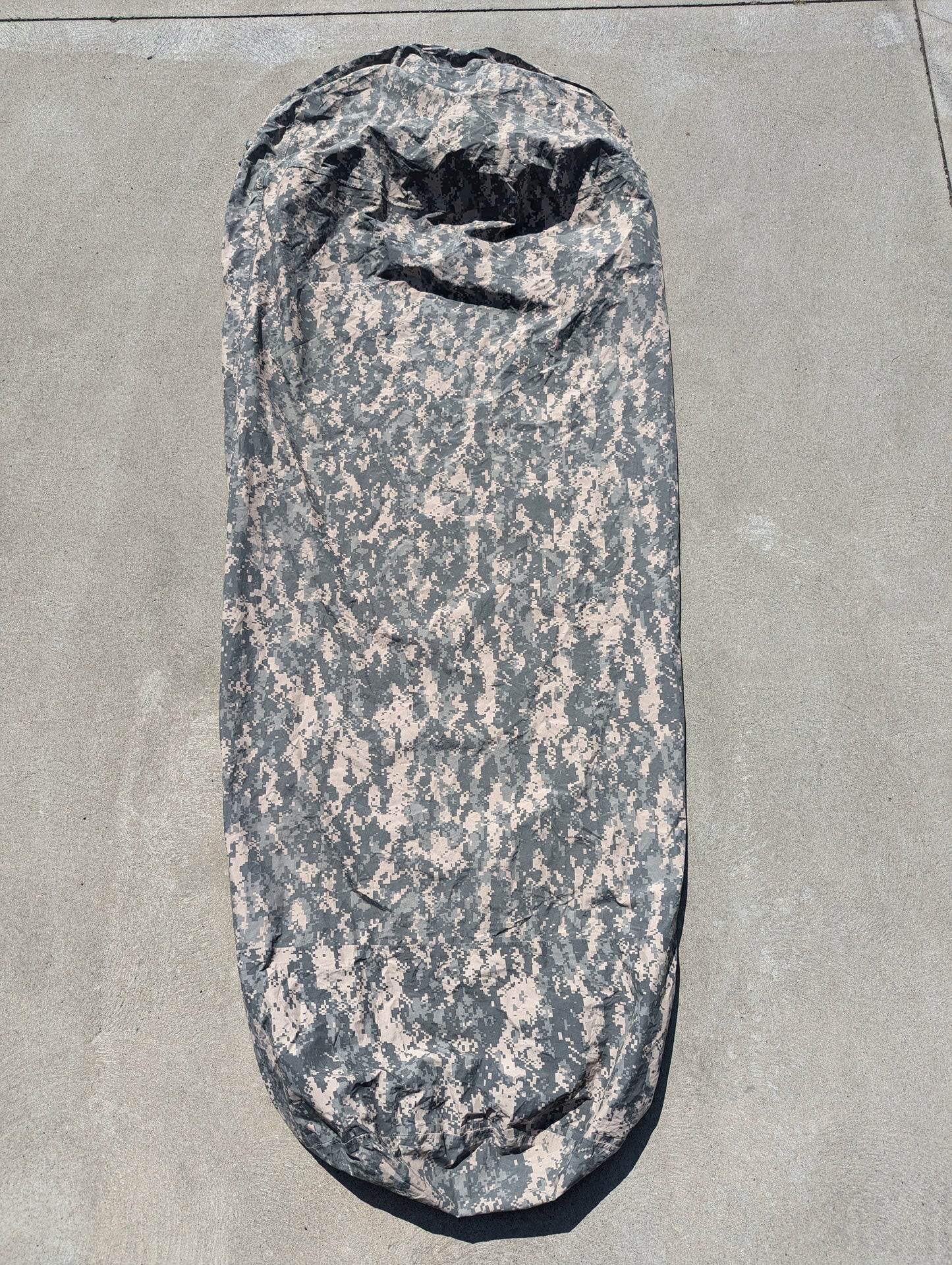 A full-length U.S. Military Surplus Modular Sleep System Bivy Cover in ACU digital camouflage, laid flat on a gray concrete surface, displaying the wrinkled mummy shape and waterproof fabric.