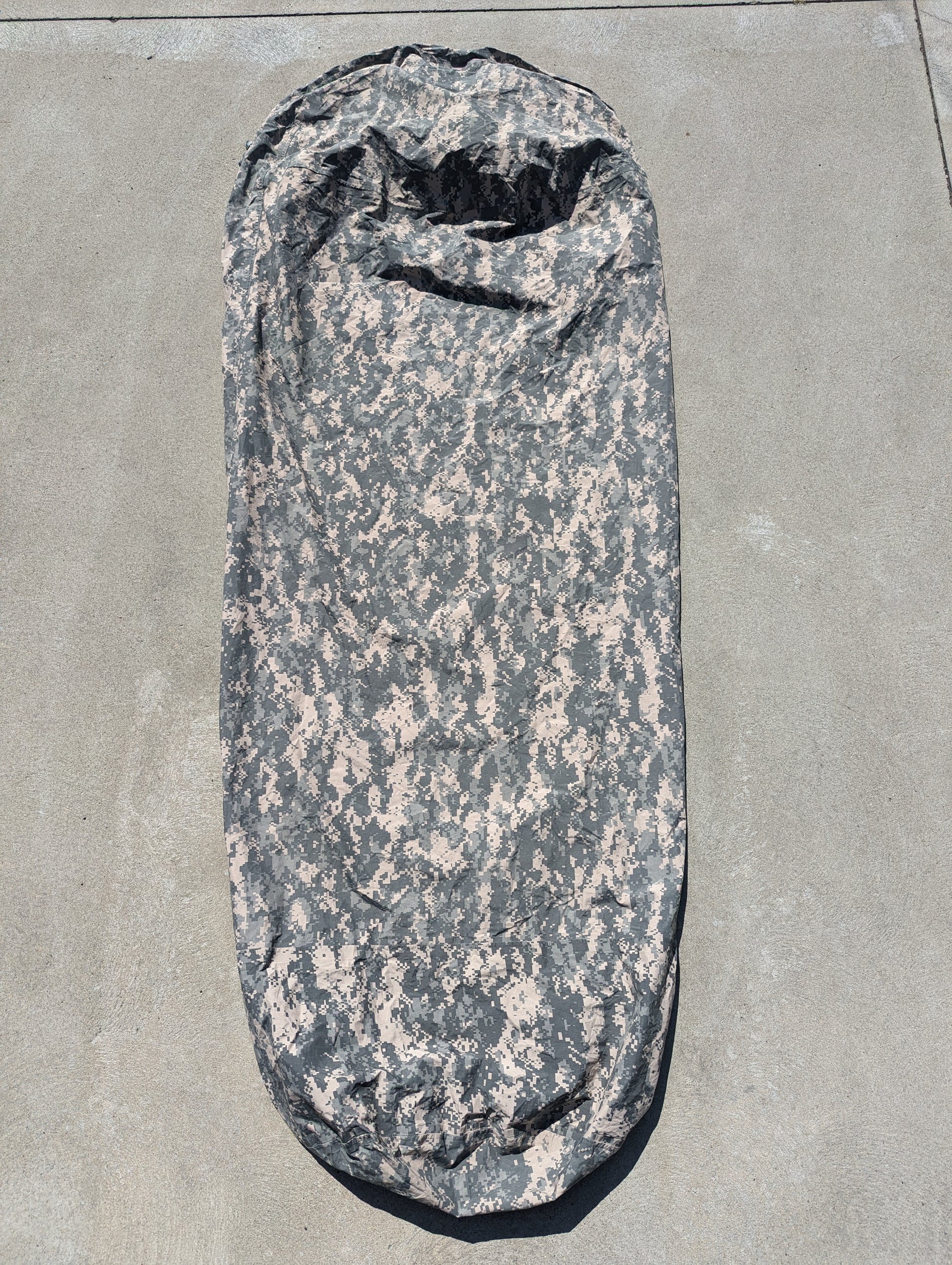 A full-length U.S. Military Surplus Modular Sleep System Bivy Cover in ACU digital camouflage, laid flat on a gray concrete surface, displaying the wrinkled mummy shape and waterproof fabric.